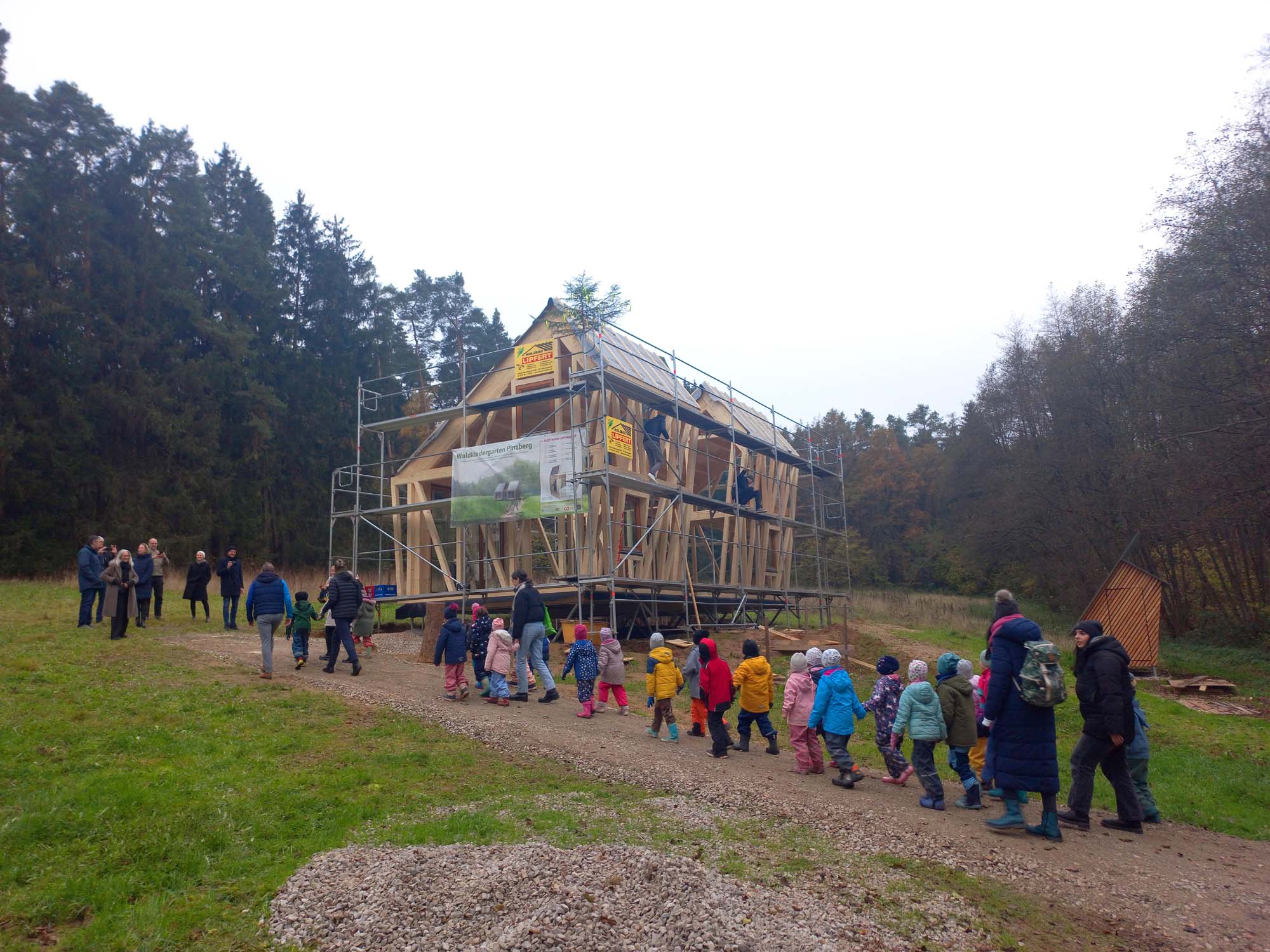 Eine Gruppe von Kindern und Erwachsenen geht auf eine im Bau befindliche Holzkonstruktion in einem grasbewachsenen Waldgebiet zu. Das Gerüst des Gebäudes ist sichtbar, im Hintergrund sind Bäume und ein bewölkter Himmel zu sehen.