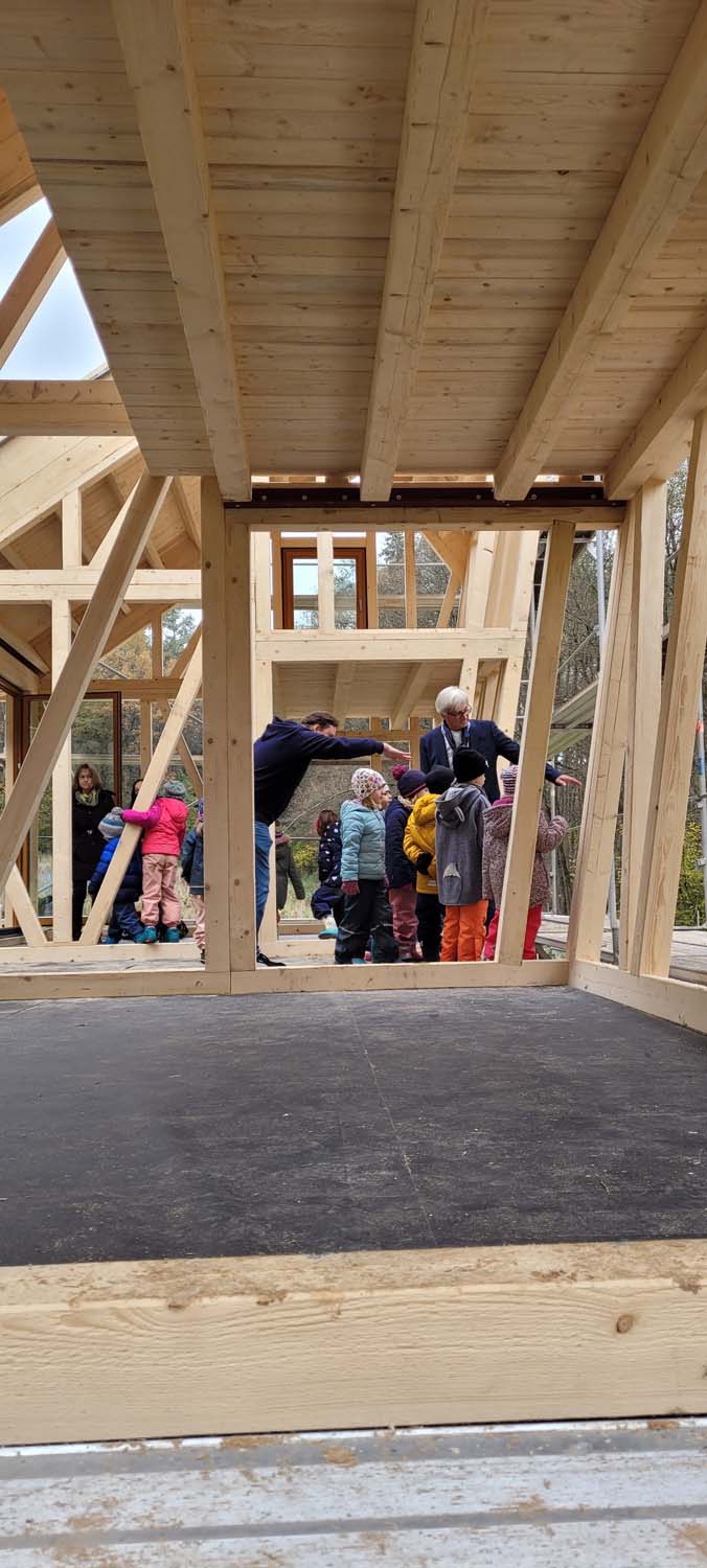 Kinder und Erwachsene versammeln sich auf einer im Bau befindlichen Holzkonstruktion, möglicherweise um an einer Gruppenaktivität oder Führung teilzunehmen. Das Gerüst umfasst Balken und offene Flächen, und die Umgebung lässt an eine Außenumgebung denken.