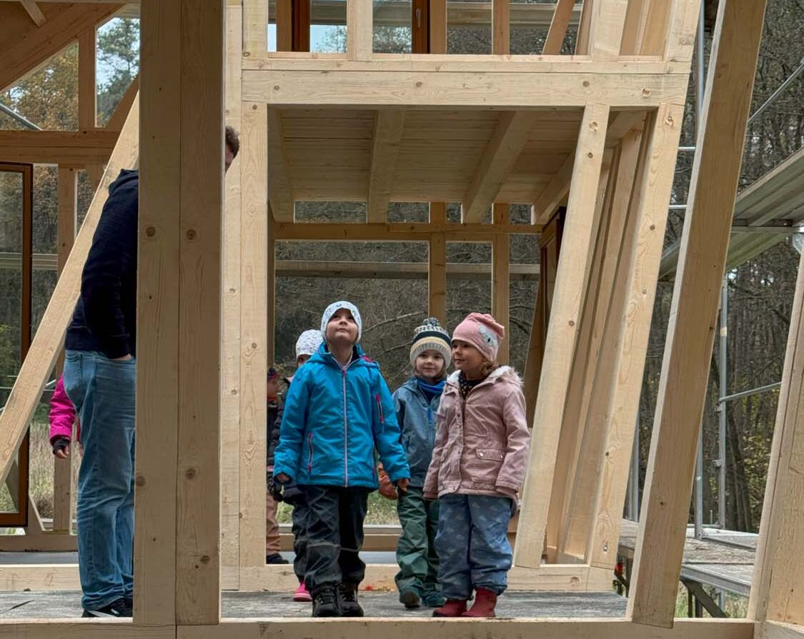 Drei Kinder stehen in einer im Bau befindlichen Holzkonstruktion und tragen warme Jacken und Mützen. Der Rahmen zeigt große Glasfenster. Auf der linken Seite steht teilweise sichtbar ein Erwachsener. Durch den offenen Rahmen sind Bäume und Himmel zu sehen.