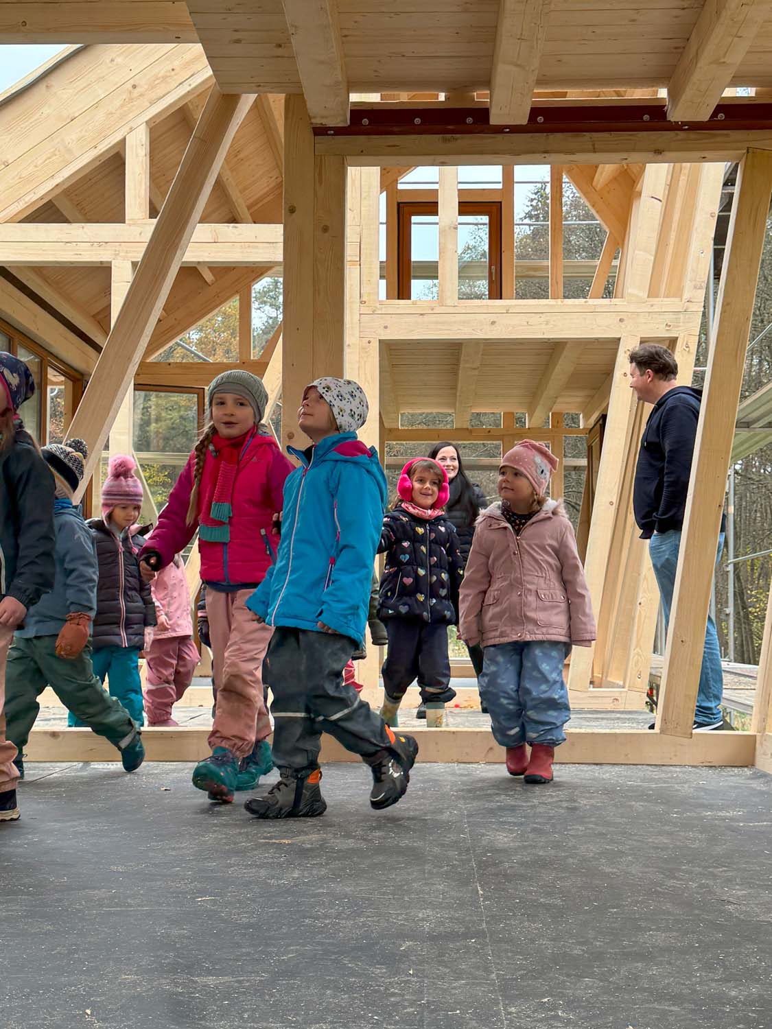 Kinder in bunter Winterkleidung gehen durch ein im Bau befindliches Holzhaus. Das Gebäude zeichnet sich durch große Balken und Fenster aus. Zwei Erwachsene stehen in der Nähe und beobachten die Szene. Im Hintergrund sind Bäume zu sehen.