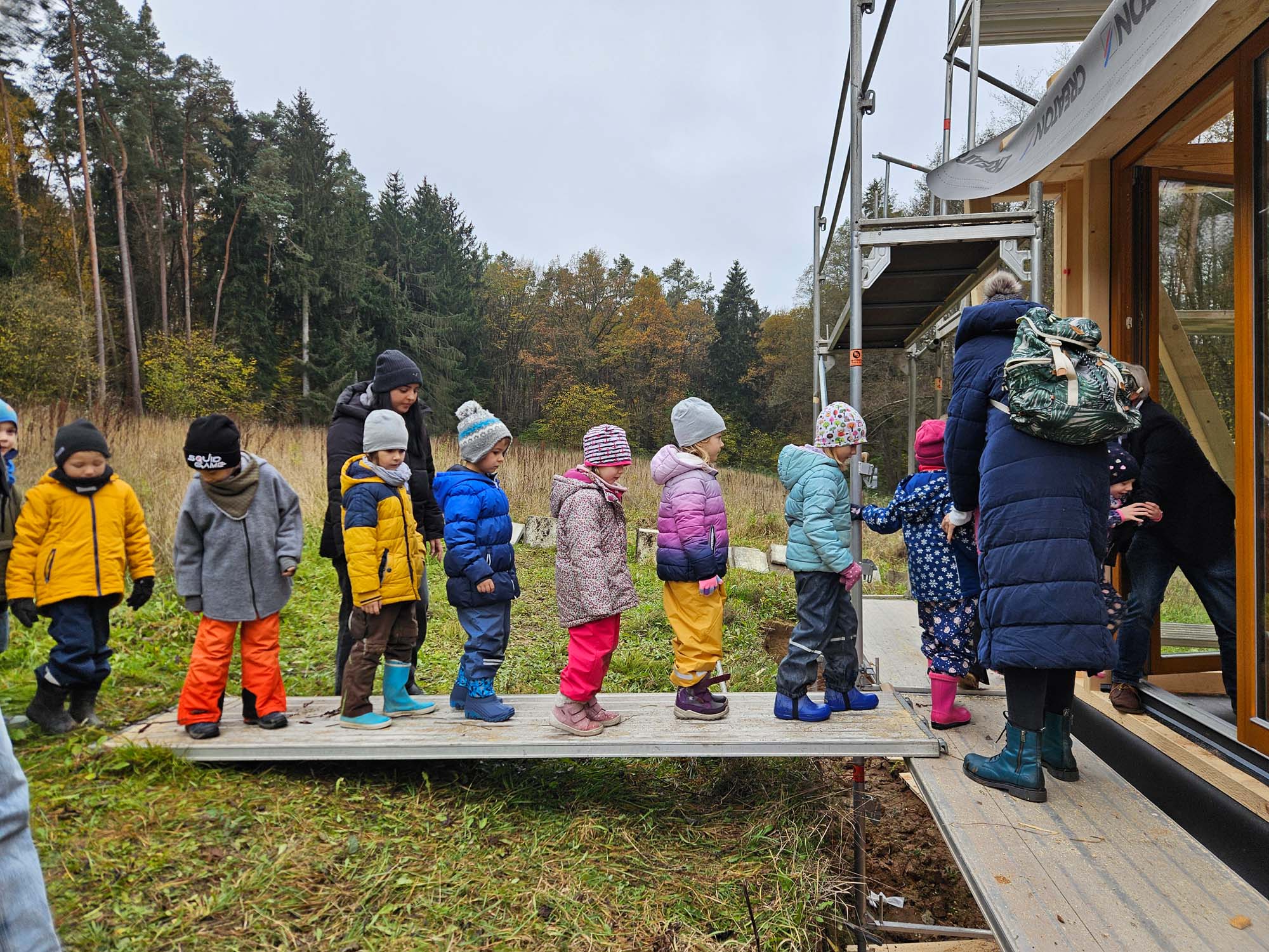 Eine Gruppe Kinder in bunter Winterkleidung steht in einer Reihe auf einer Holzrampe, die in ein Gebäude führt. Ein Erwachsener steht am Eingang und führt sie hinein. Die Szene spielt im Freien an einem bewölkten Tag mit Bäumen im Hintergrund.