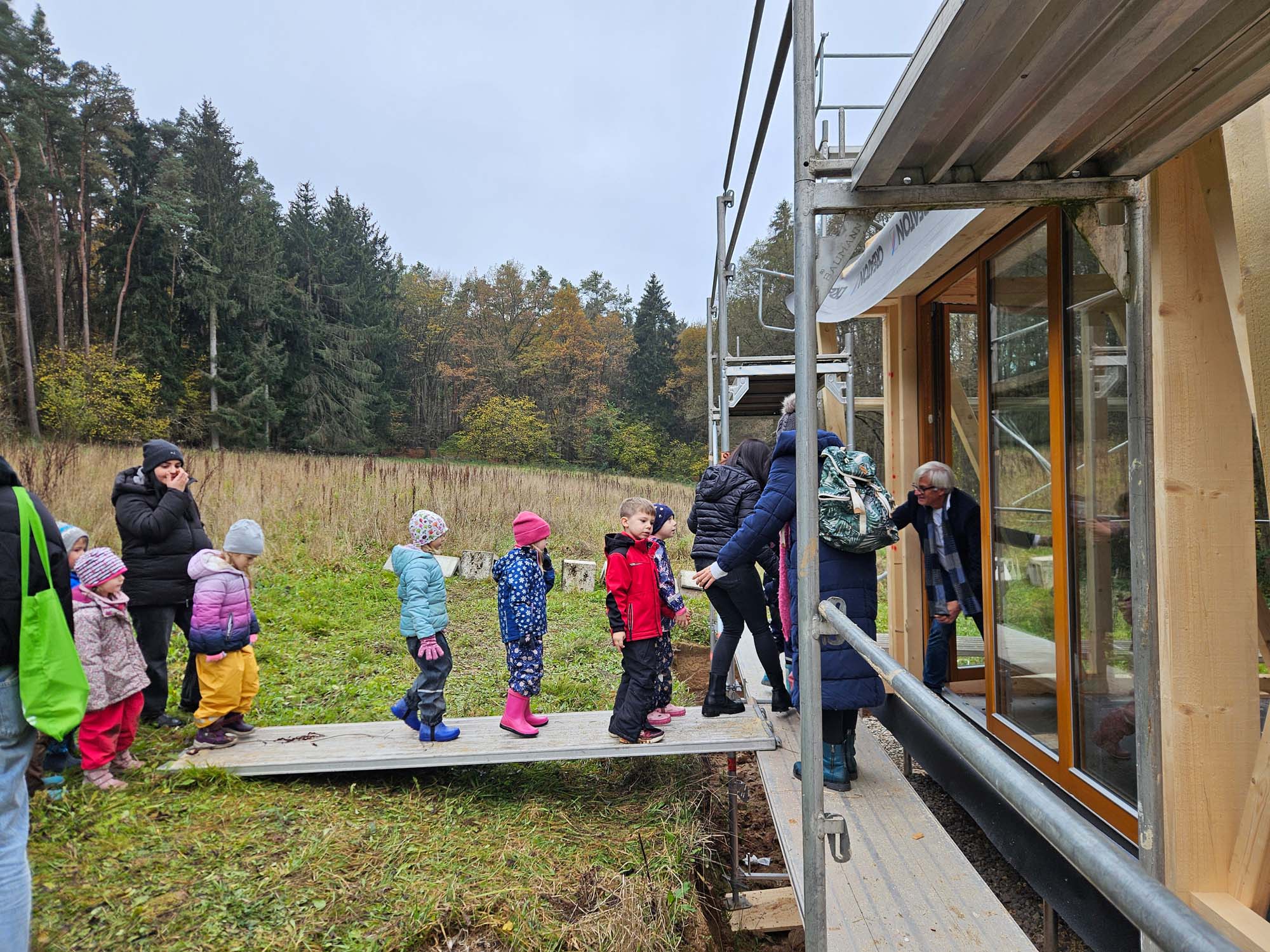 Eine Gruppe Kinder in bunten Regenmänteln und Stiefeln steht in einer Reihe auf einer Holzrampe vor einem modernen Gebäude im Bau. Erwachsene führen sie, als sie sich dem Gebäude nähern. Bäume und Gras umgeben das Gebiet unter einem bewölkten Himmel.