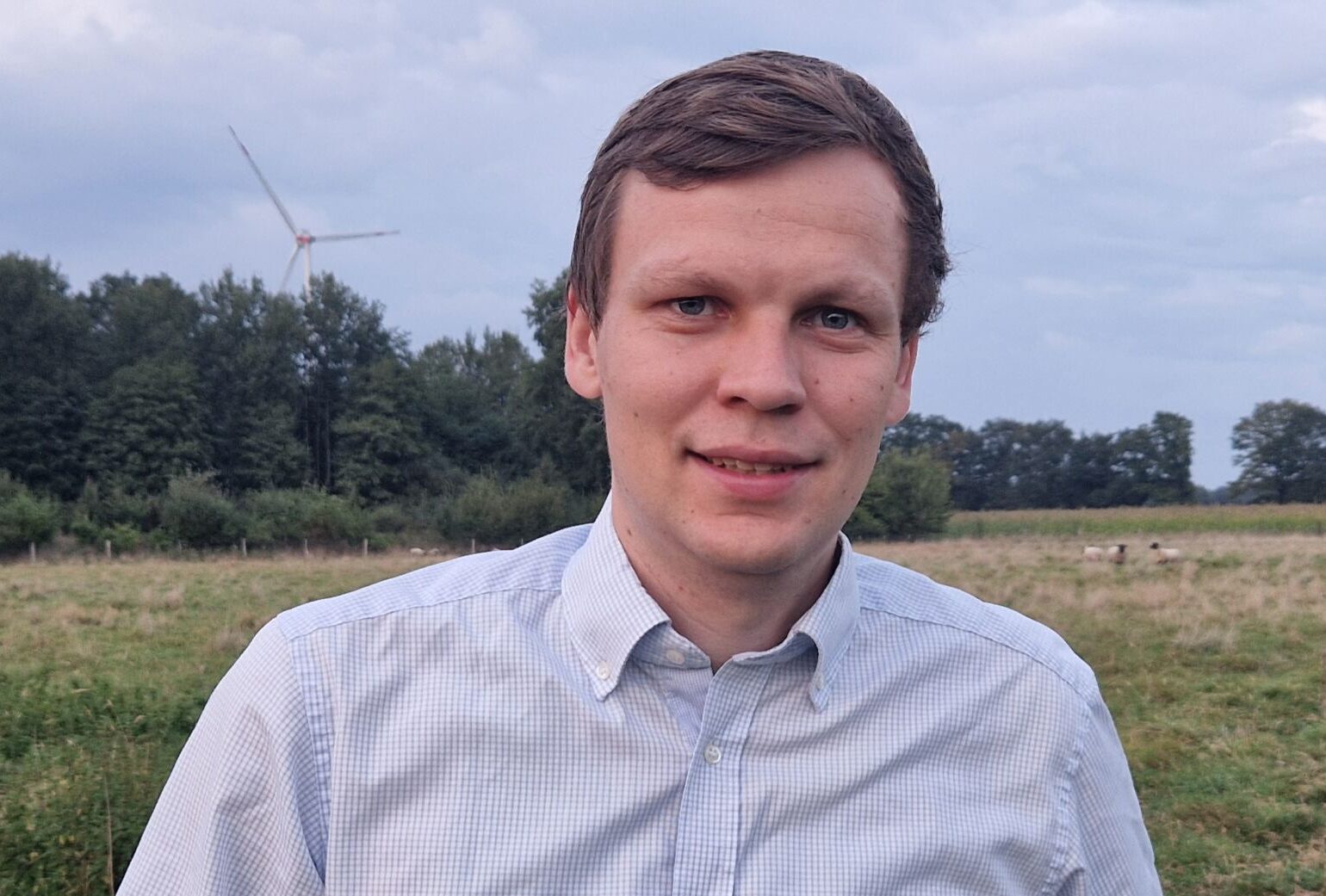 Eine Person in einem hellblauen Hemd steht unter einem bewölkten Himmel auf einer Wiese mit Bäumen und einer Windkraftanlage im Hintergrund.