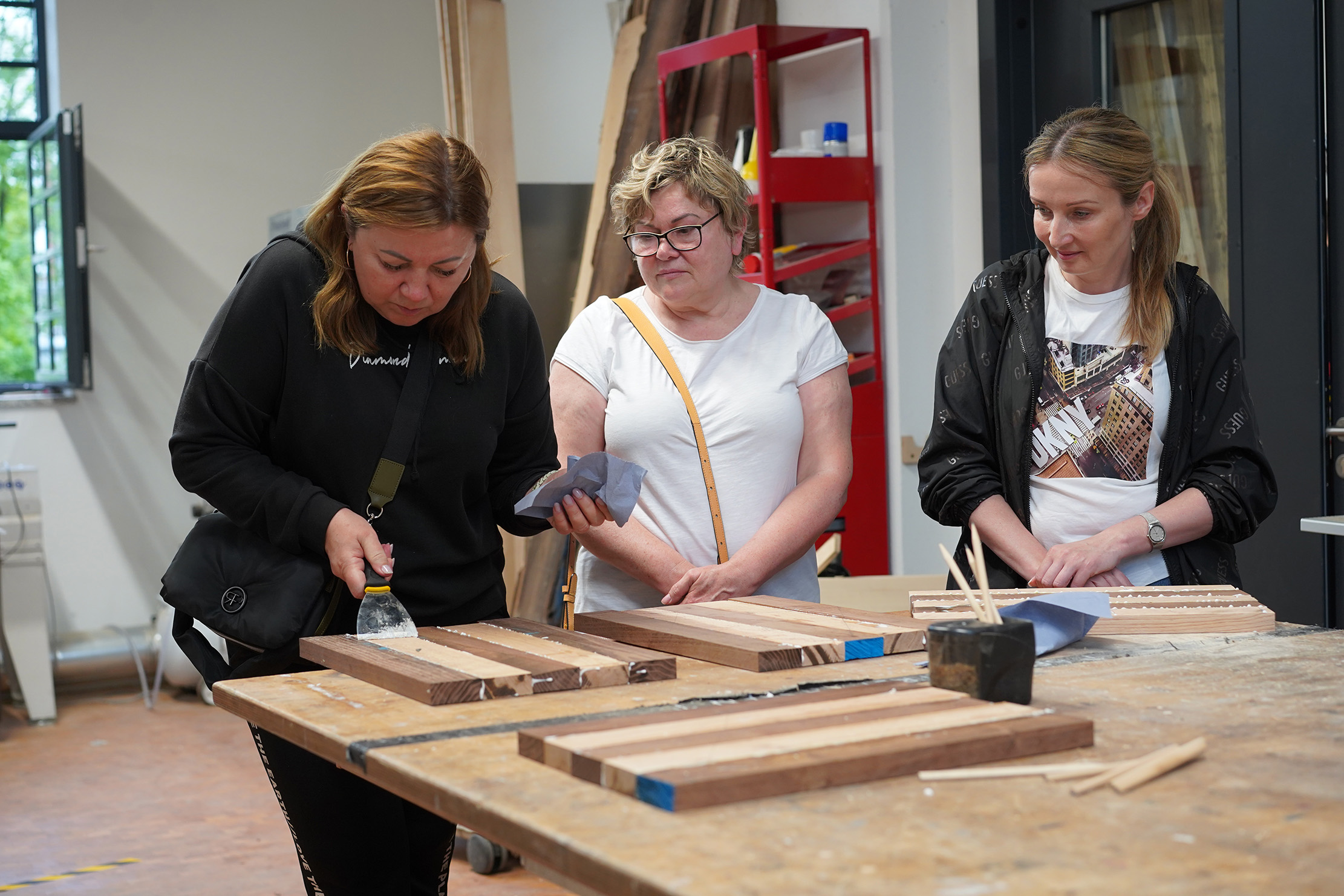 Drei Frauen arbeiten an einem Tisch in einer Werkstatt an Holzprojekten. Eine schleift ein Holzbrett, während die anderen zuschauen. Der Raum ist mit Werkzeugen und Materialien ausgestattet und durch ein Fenster fällt natürliches Licht ein.