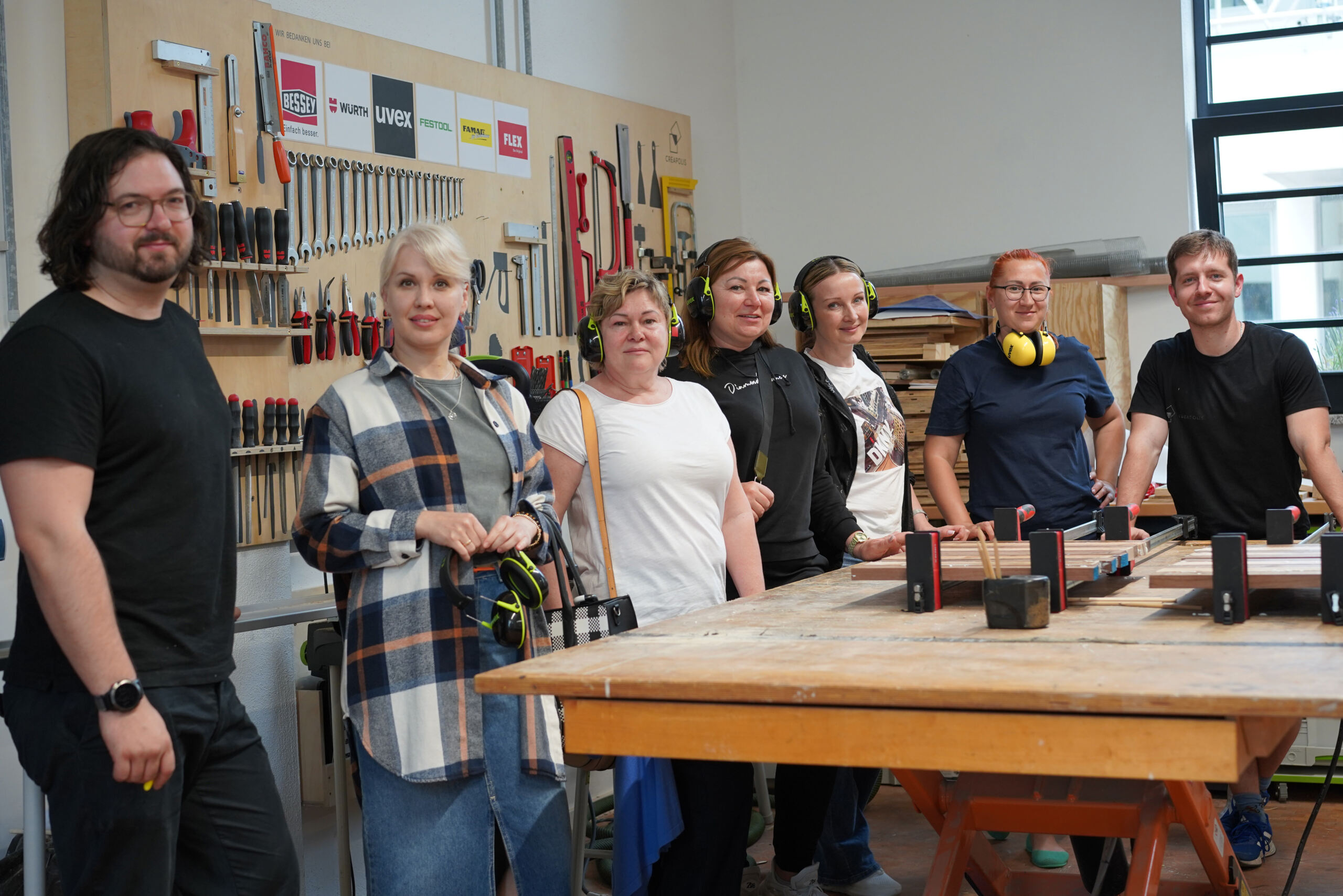 Eine Gruppe von sechs Personen steht in einer Werkstatt. An der Wand im Hintergrund hängen verschiedene Werkzeuge. Sie lächeln und blicken in die Kamera. Im Vordergrund ist eine große Holzwerkbank zu sehen. Einige Personen tragen Freizeitkleidung und auf der Werkbank liegen Kopfhörer.