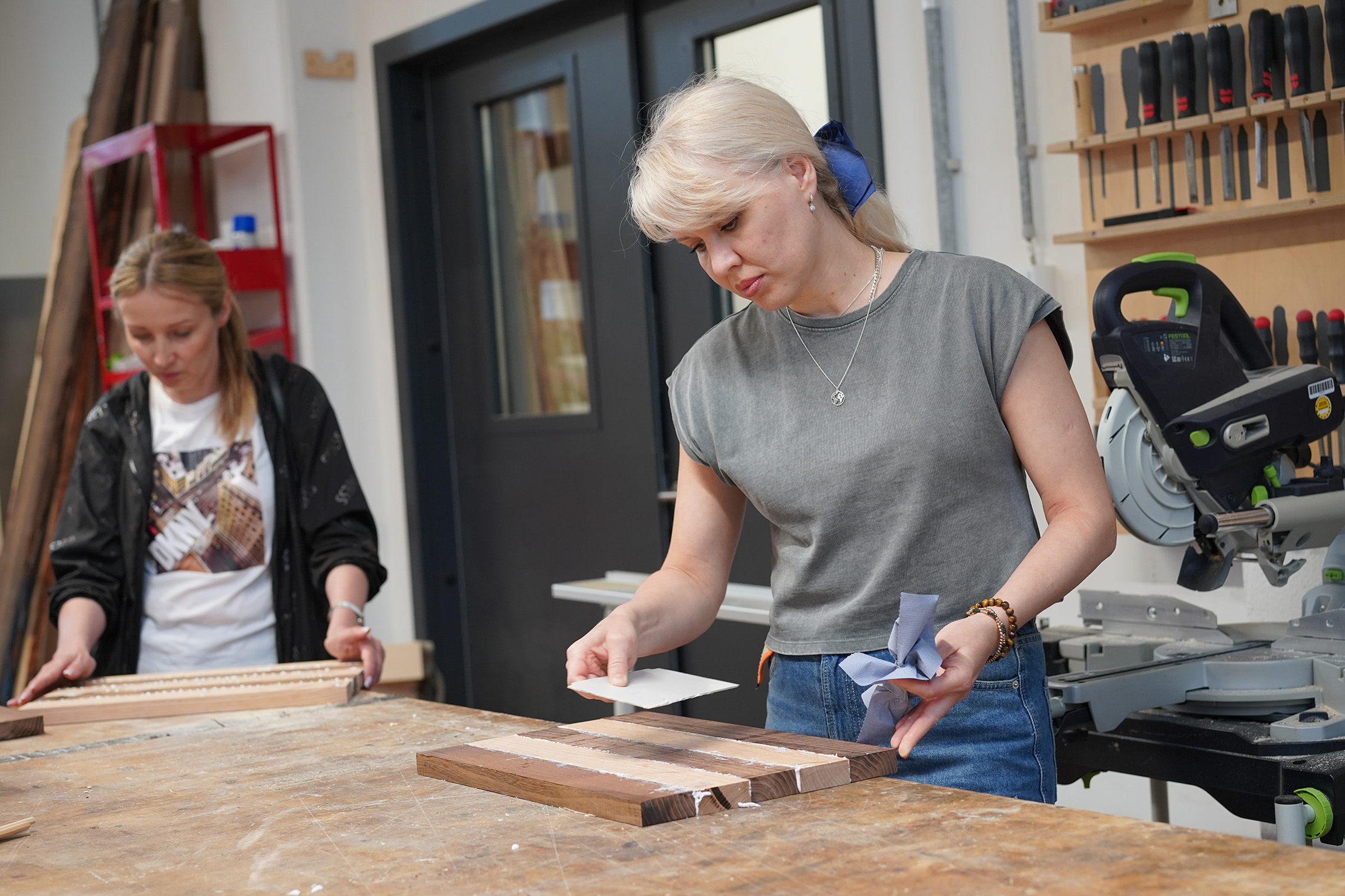 Zwei Frauen arbeiten in einer Tischlerwerkstatt. Eine schleift ein Stück Holz, während die andere ein anderes Stück vorbereitet. Um sie herum sind verschiedene Werkzeuge und Materialien zu sehen, darunter eine Säge und Regale mit Werkzeugen an der Wand.