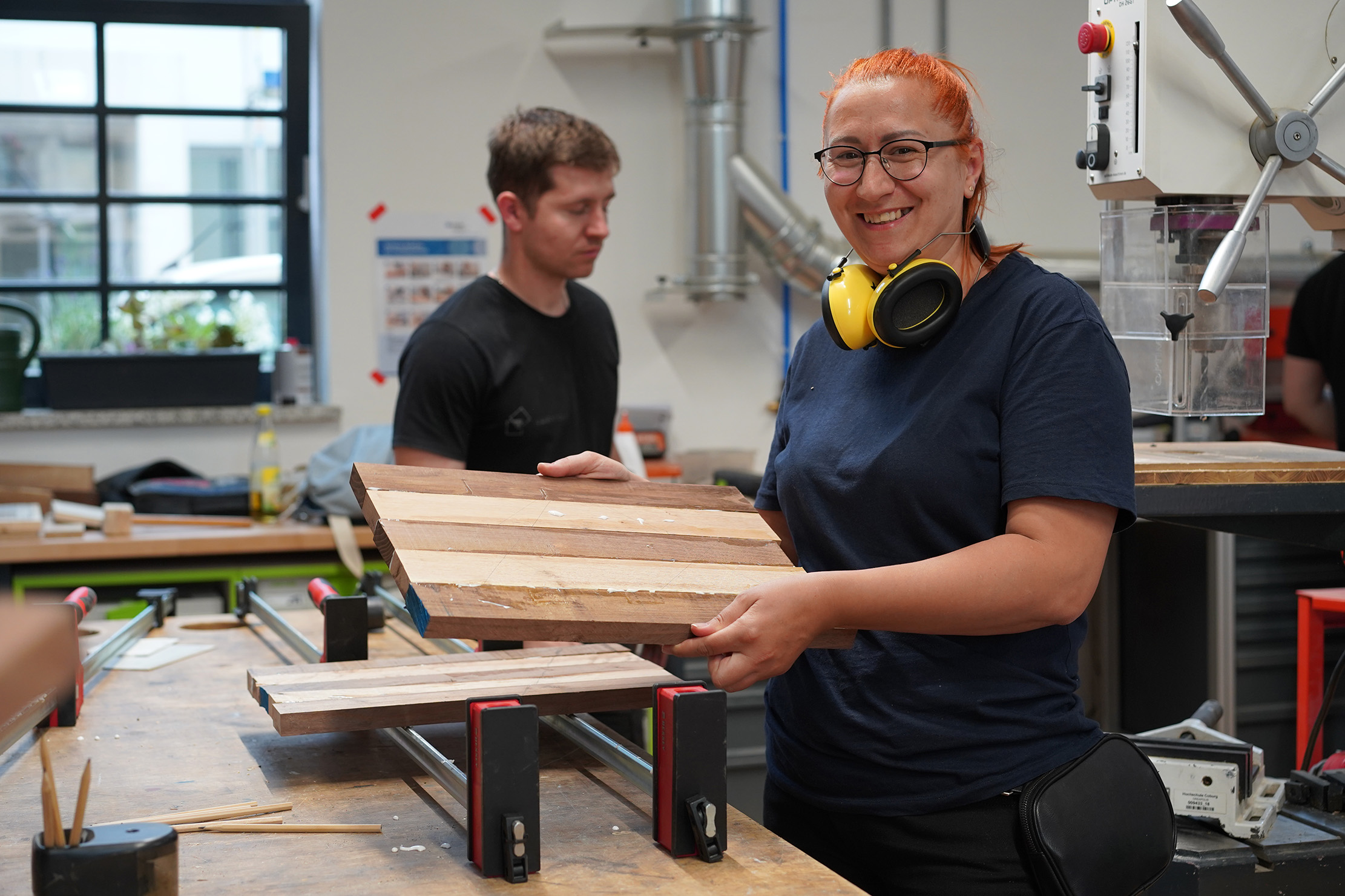 Eine Frau lächelt und hält ein Holzbrett in einer Holzwerkstatt. Sie trägt Sicherheitskopfhörer um den Hals. Im Hintergrund arbeitet ein Mann an einem Tisch mit verschiedenen Werkzeugen und Holzbrettern. Durch das große Fenster fällt natürliches Licht ein.