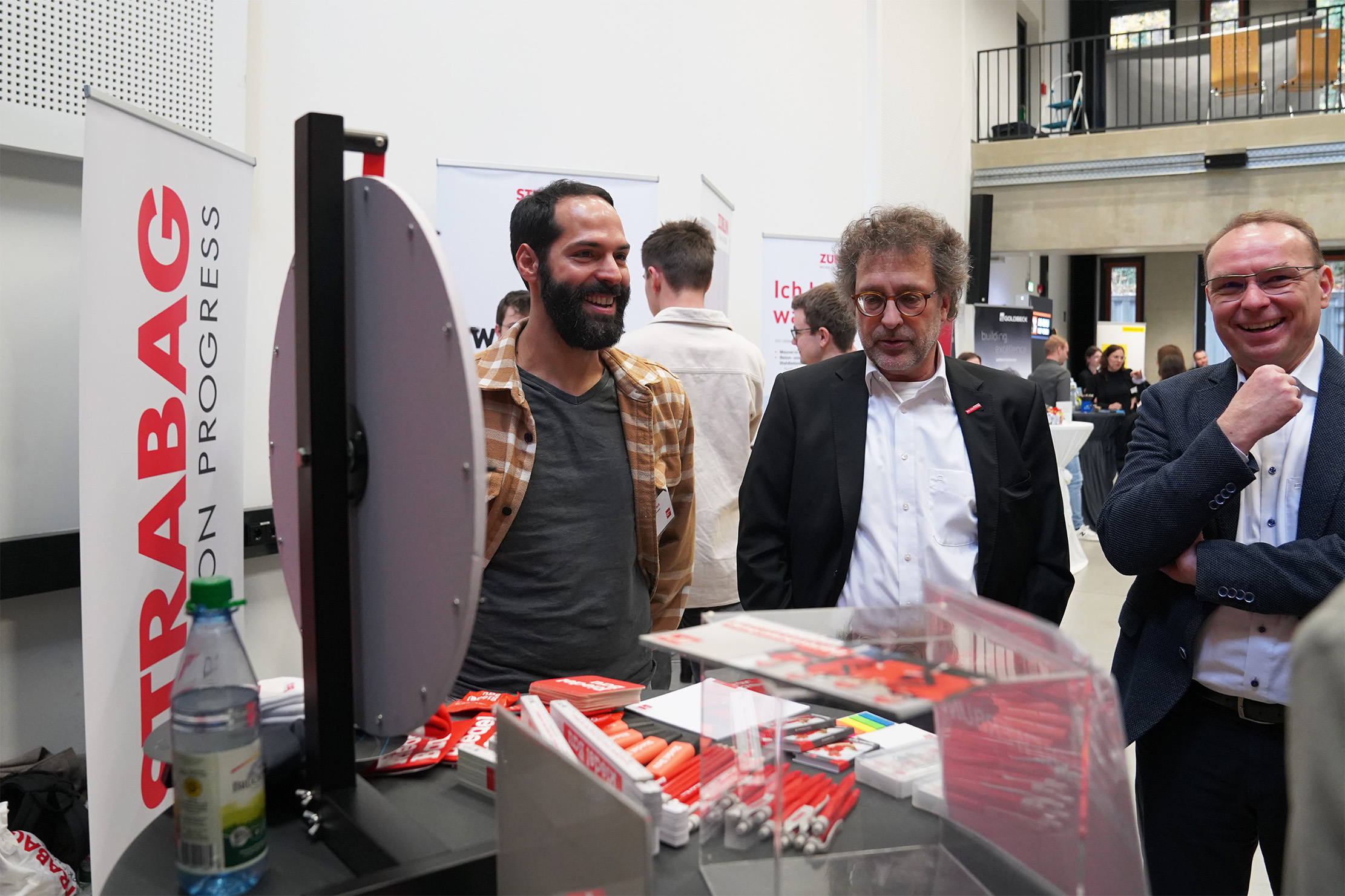 Drei Männer stehen bei einer Strabag-Veranstaltung neben einem Tisch mit Werbematerialien. Auf einem Display sind das Strabag-Logo und der Slogan „ON PROGRESS“ zu sehen. Im Hintergrund sind weitere Teilnehmer und Eventdisplays zu sehen.