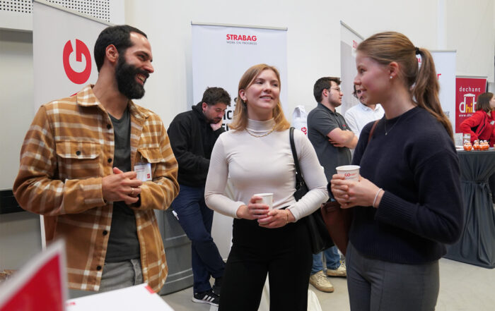 Drei Menschen unterhalten sich fröhlich bei einer Veranstaltung. Ein Mann im karierten Hemd hält ein Schlüsselband, während zwei Frauen, eine davon mit einer Tasse, zuhören und lächeln. Im Hintergrund unterhalten sich mehrere andere an Ständen mit Bannern.