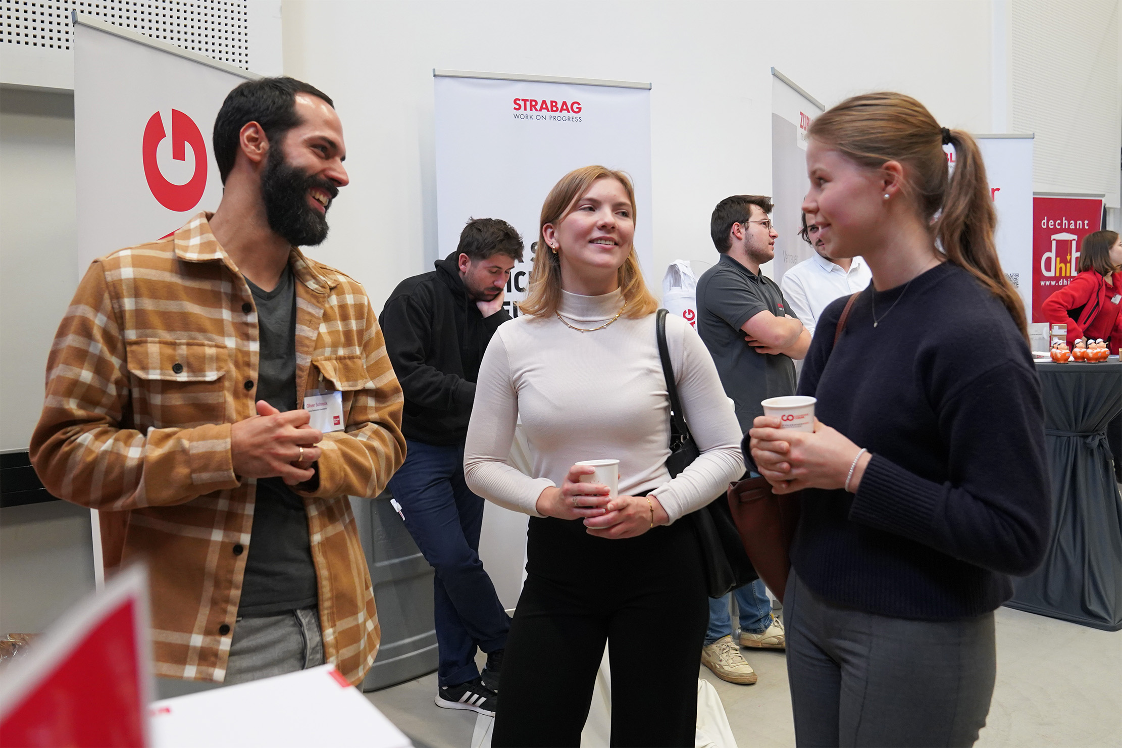 Drei Menschen unterhalten sich fröhlich bei einer Veranstaltung. Ein Mann im karierten Hemd hält ein Schlüsselband, während zwei Frauen, eine davon mit einer Tasse, zuhören und lächeln. Im Hintergrund unterhalten sich mehrere andere an Ständen mit Bannern.