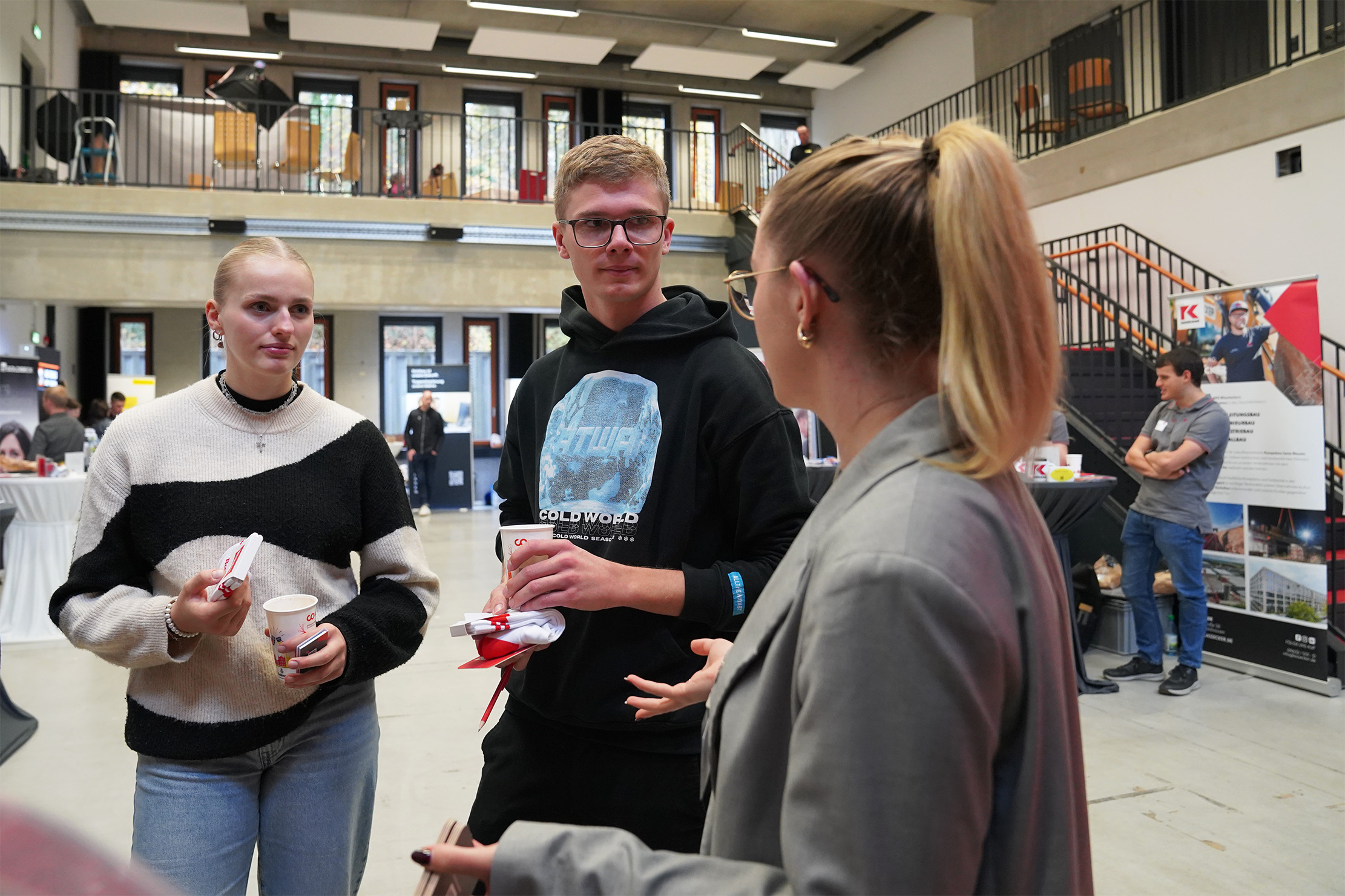 Drei Personen unterhalten sich bei einer Indoor-Veranstaltung. Eine Person hält ein Getränk und zwei haben Snacks in der Hand. Im Hintergrund sind ein Zwischengeschoss, Veranstaltungsstände und eine stehende Person neben einem Banner zu sehen.