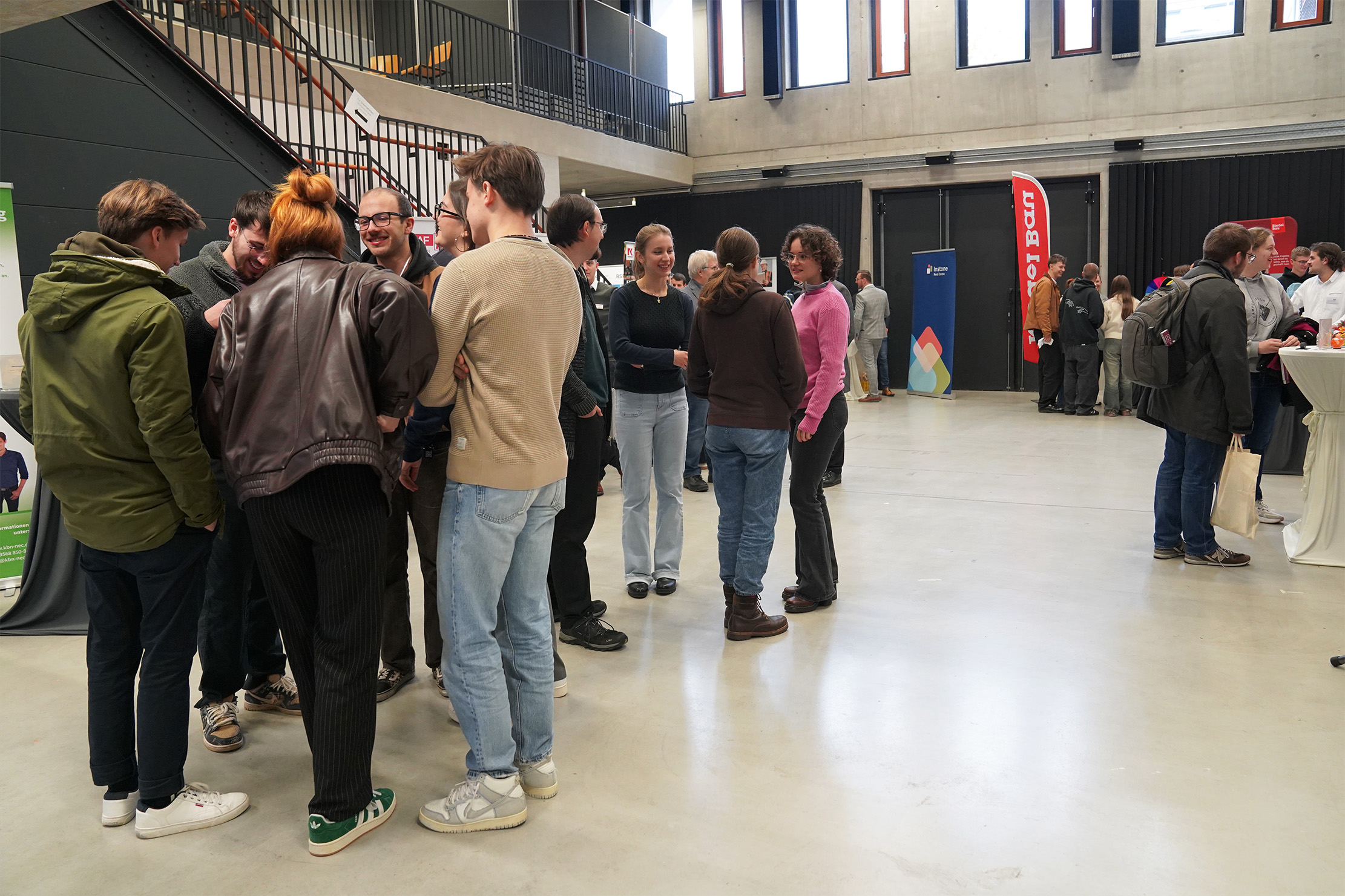 Bei einer Indoor-Veranstaltung mit Informationsständen und Tischen treffen sich Menschen in Gruppen und unterhalten sich. Der Raum zeichnet sich durch hohe Decken und moderne Architektur aus, durch die Fenster strömt natürliches Licht.