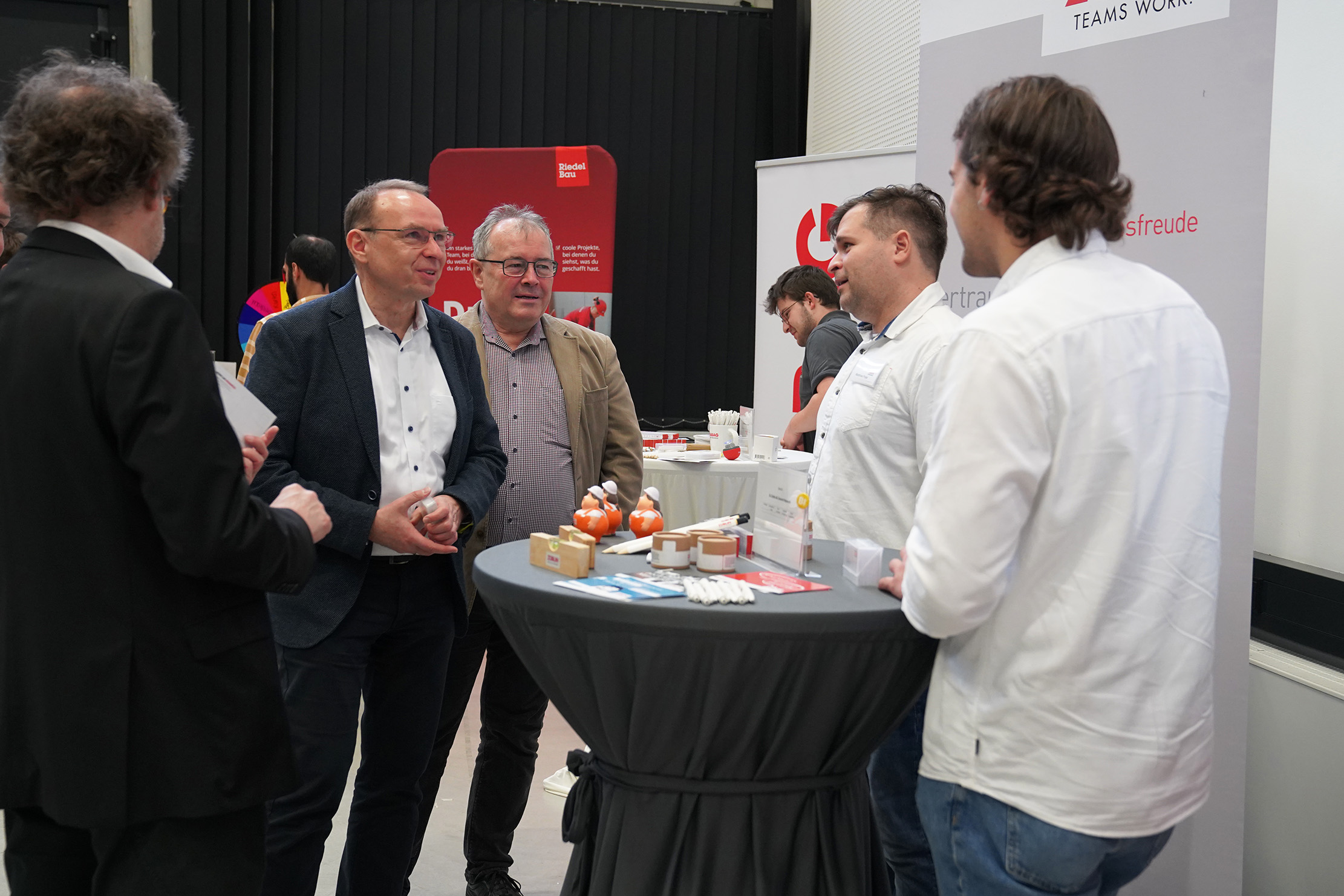 Bei einer Veranstaltung steht eine Gruppe von Menschen um einen hohen Tisch. Einige halten Broschüren und Visitenkarten in den Händen. Im Hintergrund sind Ausstellungsständer und ein Banner mit einem Logo zu sehen. Die Atmosphäre ist professionell und gesprächig.