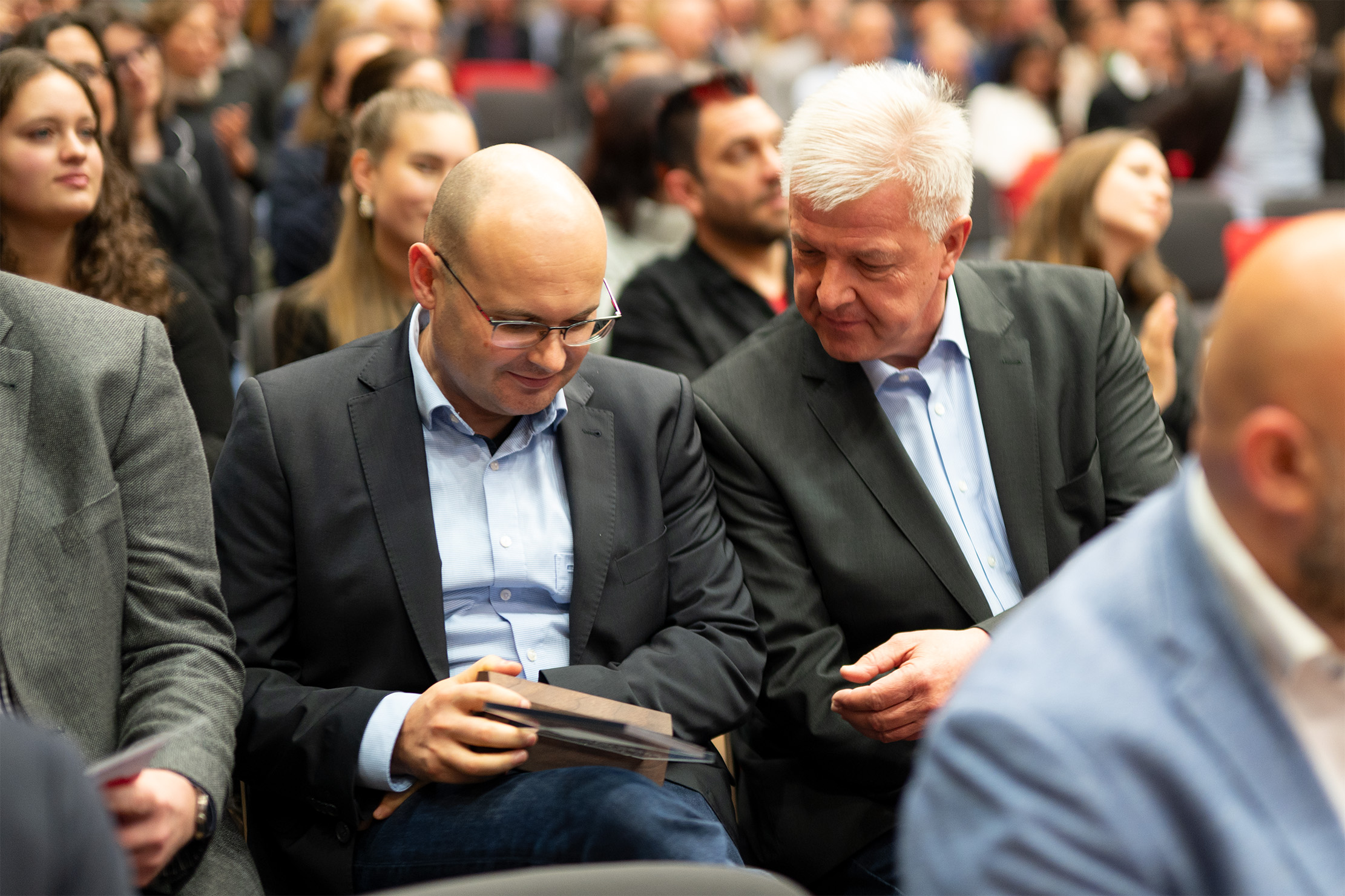 Zwei Männer in Anzügen sitzen in einem Auditorium inmitten einer Menschenmenge. Einer der Männer hält ein Buch in der Hand und scheint in ein Gespräch mit dem anderen vertieft zu sein. Das Publikum hinter ihnen sitzt und schaut aufmerksam zu.