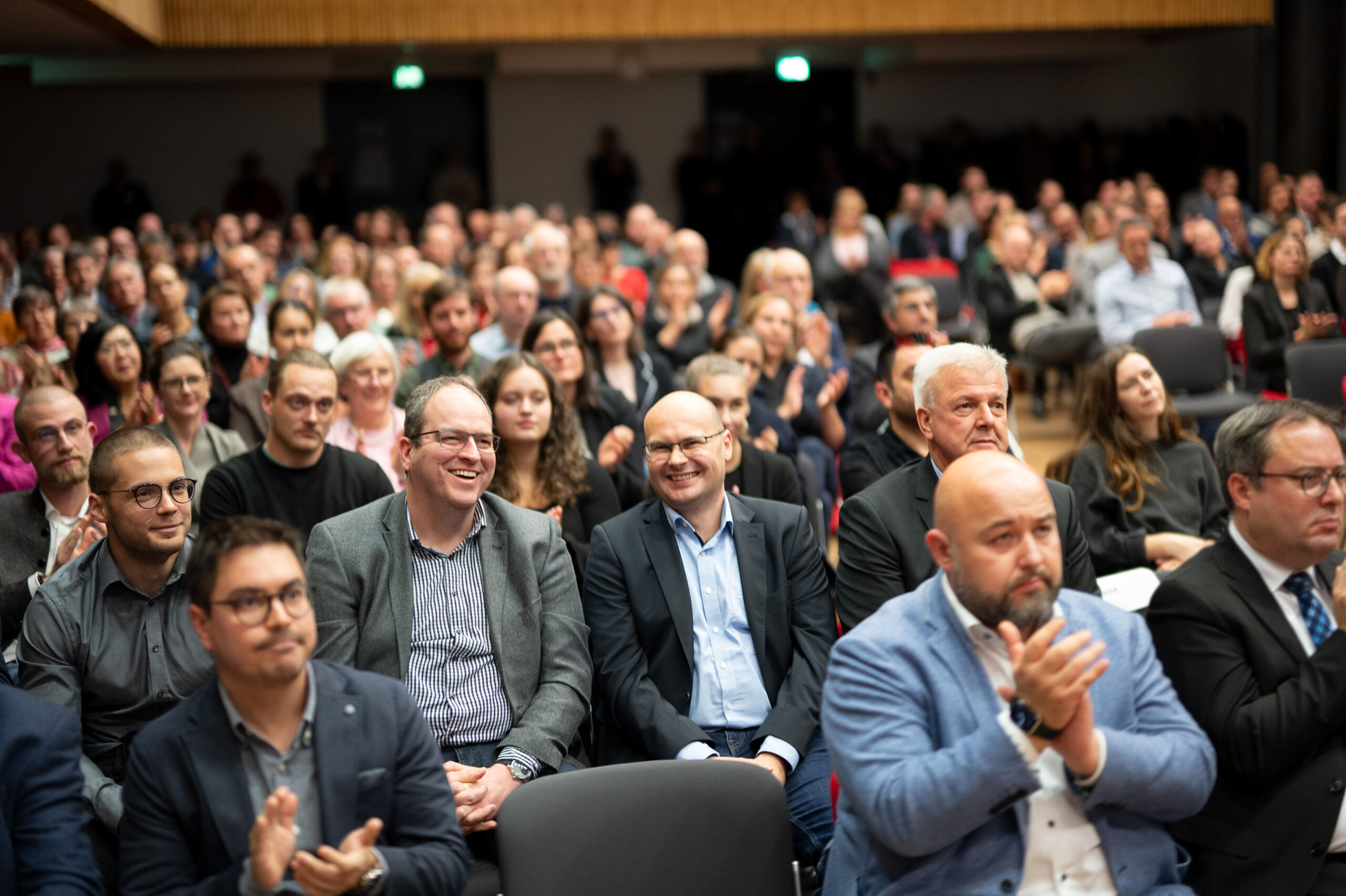 Eine große Gruppe von Menschen sitzt bei einer Konferenz oder Veranstaltung in Reihen, einige klatschen und lächeln. Das Publikum ist gemischten Geschlechts und Alters und befindet sich in einem gut beleuchteten Auditorium mit Holzakzenten.