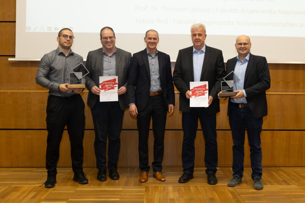 Fünf Männer stehen auf einer Holzbühne, vier halten Auszeichnungen und zwei Zertifikate in den Händen. Sie sind in Business-Kleidung gekleidet, lächeln und hinter ihnen ist ein Präsentationsbildschirm mit einem deutschen Text zu sehen.