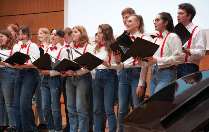 Auf der Bühne singt ein Chor junger Leute in weißen Hemden, roten Hosenträgern und roten Fliegen. Sie halten schwarze Mappen in den Händen und stehen vor einer Holzkulisse, im Vordergrund steht ein Klavier.