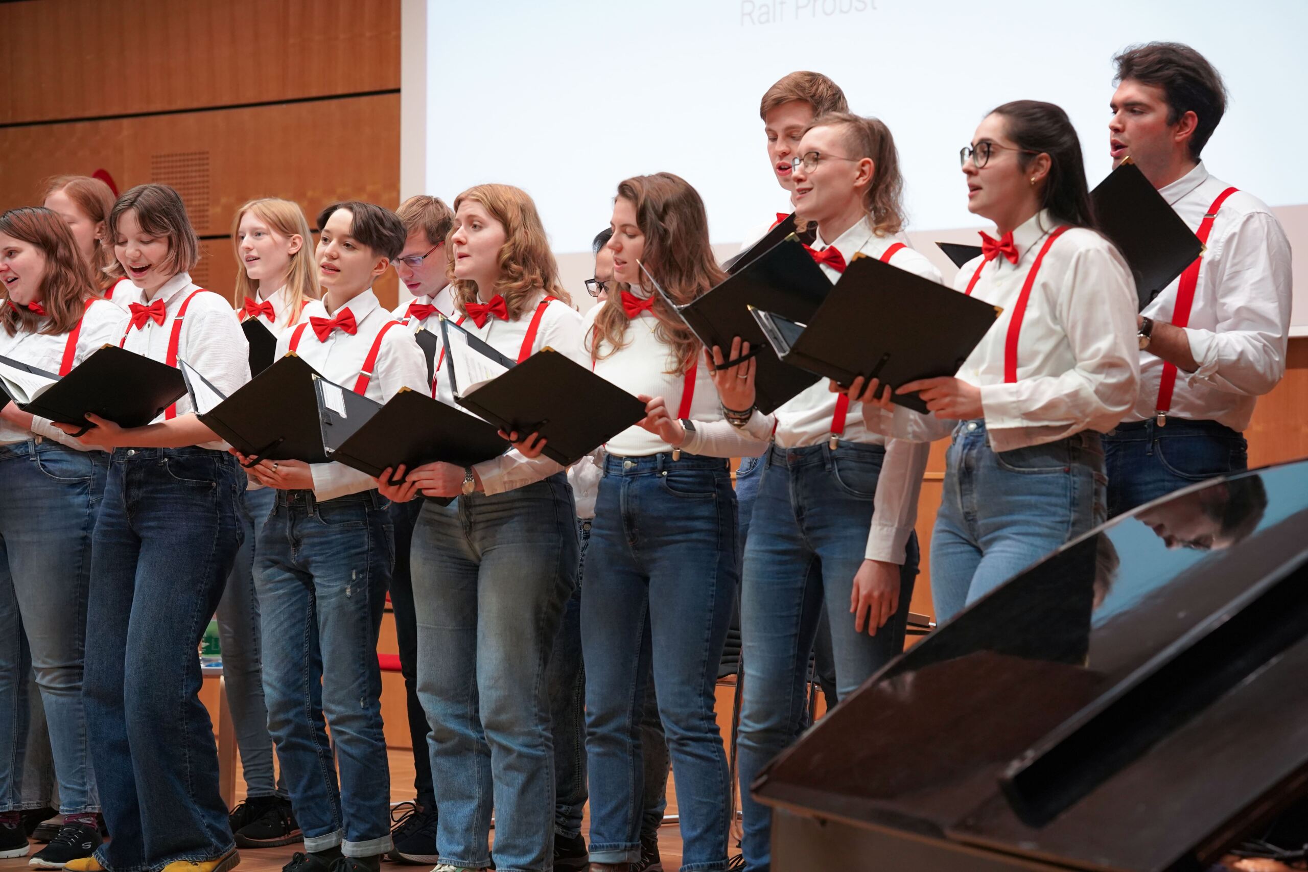 Auf der Bühne singt ein Chor junger Leute in weißen Hemden, roten Hosenträgern und roten Fliegen. Sie halten schwarze Mappen in den Händen und stehen vor einer Holzkulisse, im Vordergrund steht ein Klavier.