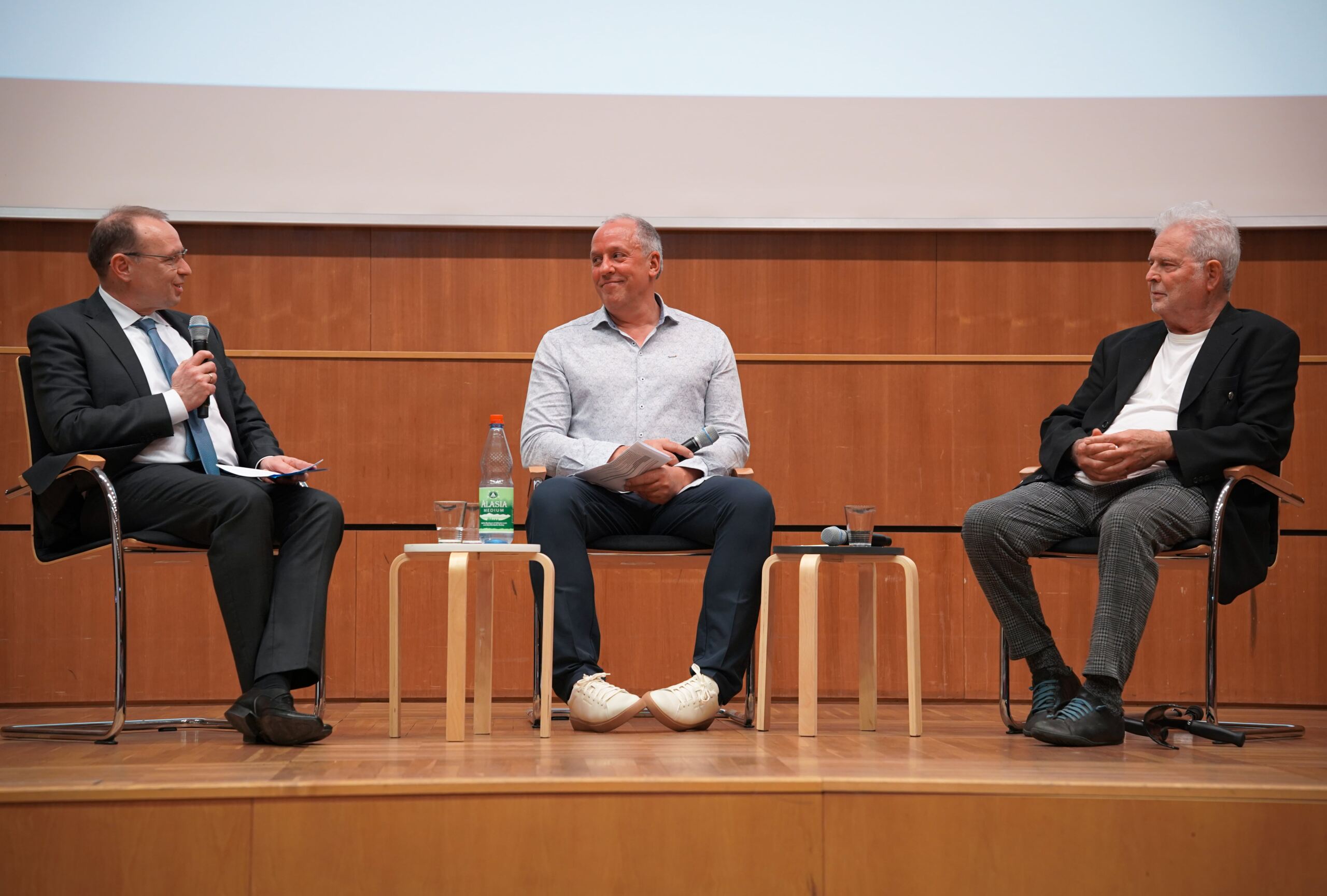 Drei Männer sitzen bei einer Podiumsdiskussion auf einer Bühne. Einer hält ein Mikrofon und stellt Fragen. Auf kleinen Tischen zwischen ihnen stehen eine Flasche Wasser und Gläser. Im Hintergrund ist eine Holzwand zu sehen, über der eine große Leinwand hängt.