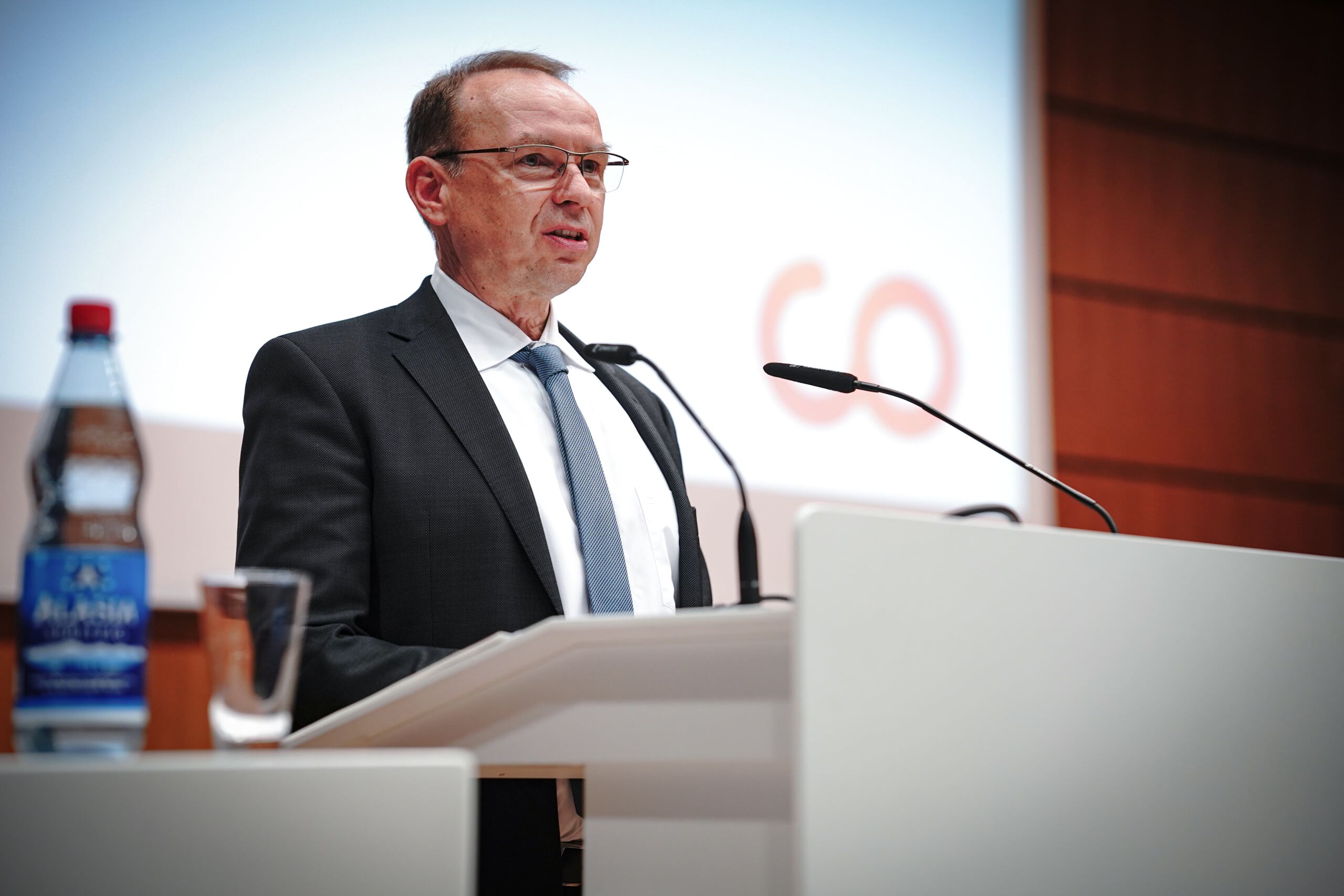 Ein Mann im Anzug spricht an einem Podium mit mehreren Mikrofonen. Auf dem Schreibtisch stehen eine Wasserflasche und ein Glas. Im Hintergrund ist ein unscharfer Bildschirm zu sehen, auf dem Teile der Buchstaben zu sehen sind.