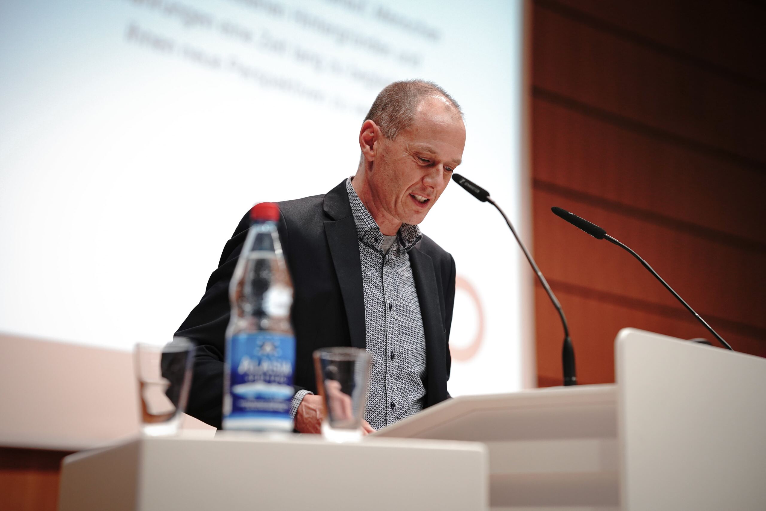 Eine Person im Anzug spricht an einem Podium mit Mikrofonen. Auf dem Podium stehen eine Flasche Wasser und ein Glas. Im Hintergrund ist eine Projektion mit teilweise sichtbarem Text zu sehen. Die Kulisse scheint ein Konferenz- oder Hörsaal zu sein.