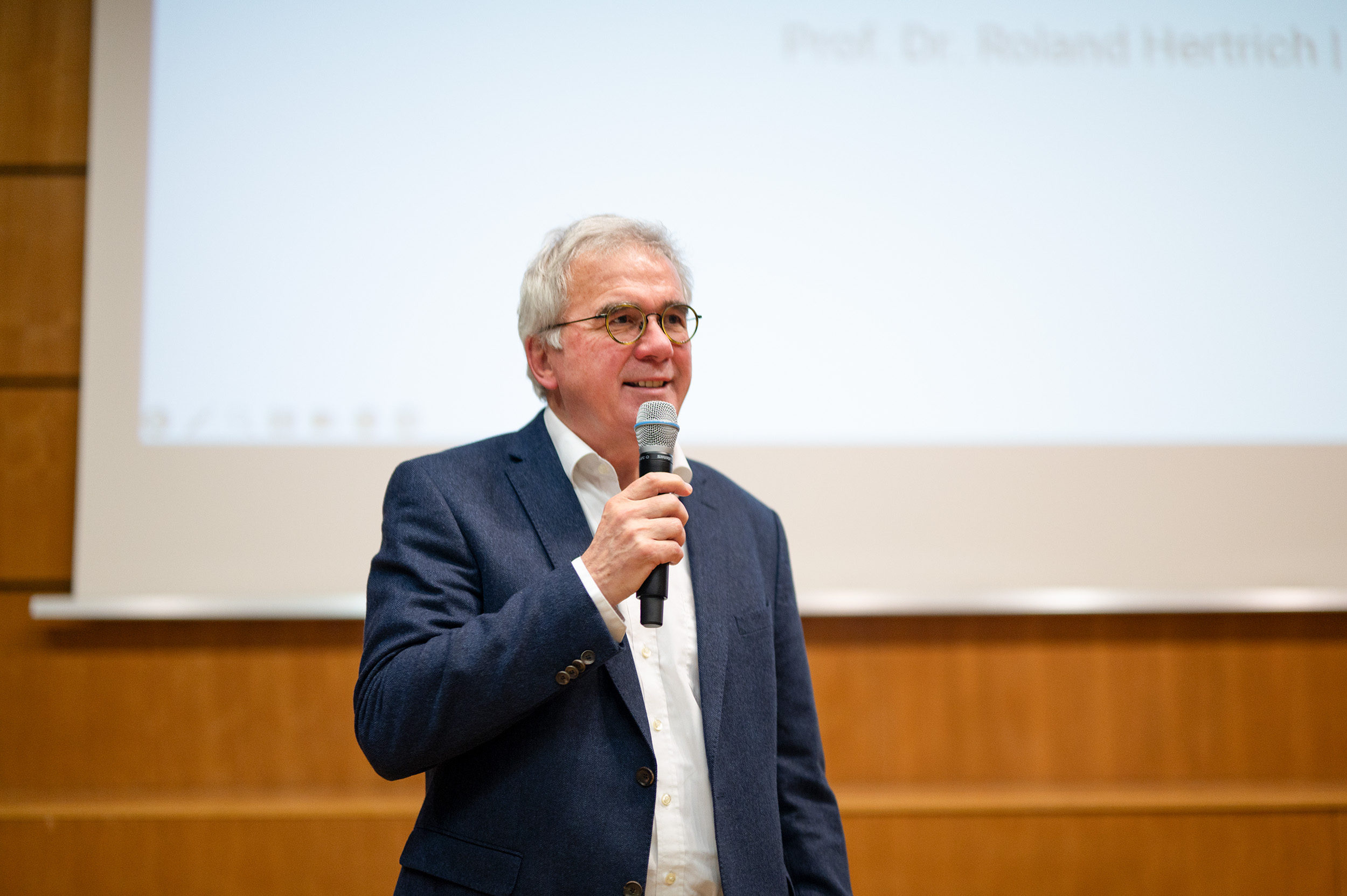 Ein älterer Mann mit grauem Haar und Brille, der einen blauen Blazer und ein weißes Hemd trägt, hält ein Mikrofon und steht vor einem weißen Bildschirm in einem Hörsaal. Der Hintergrund ist eine Holzvertäfelung.