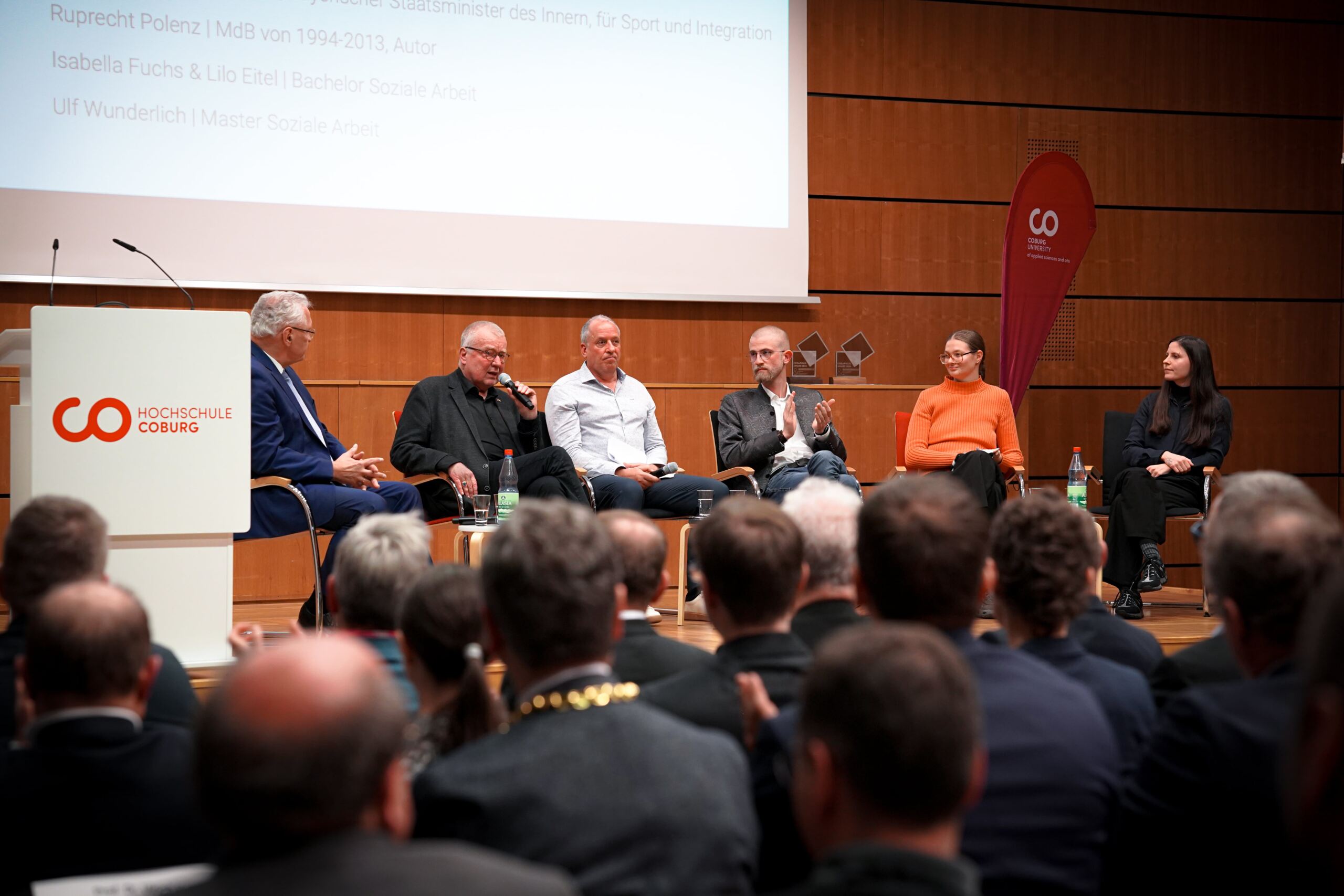 Ein sechsköpfiges Gremium sitzt bei einer Diskussion an der Hochschule Coburg auf der Bühne. Sie sitzen auf Stühlen mit Mikrofonen, während das Publikum zusieht. Auf der Bühne sind ein Banner und ein Podium zu sehen.