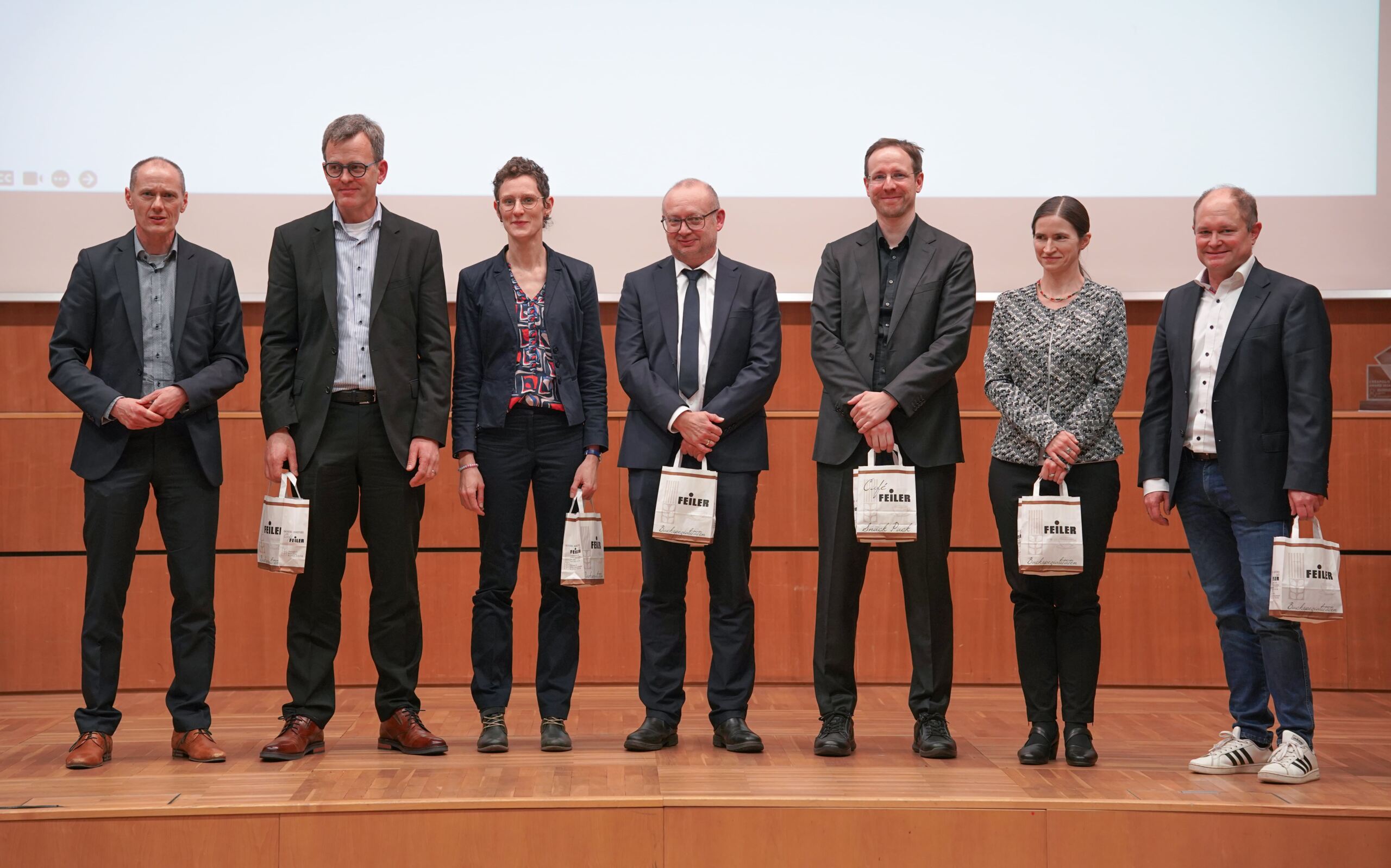 Sieben Personen stehen auf einer Bühne und halten jeweils eine kleine Geschenktüte in der Hand. Sie sind in formelle und halbformelle Kleidung gekleidet und stehen vor einem hölzernen Hintergrund mit einer leeren Leinwand über ihnen.