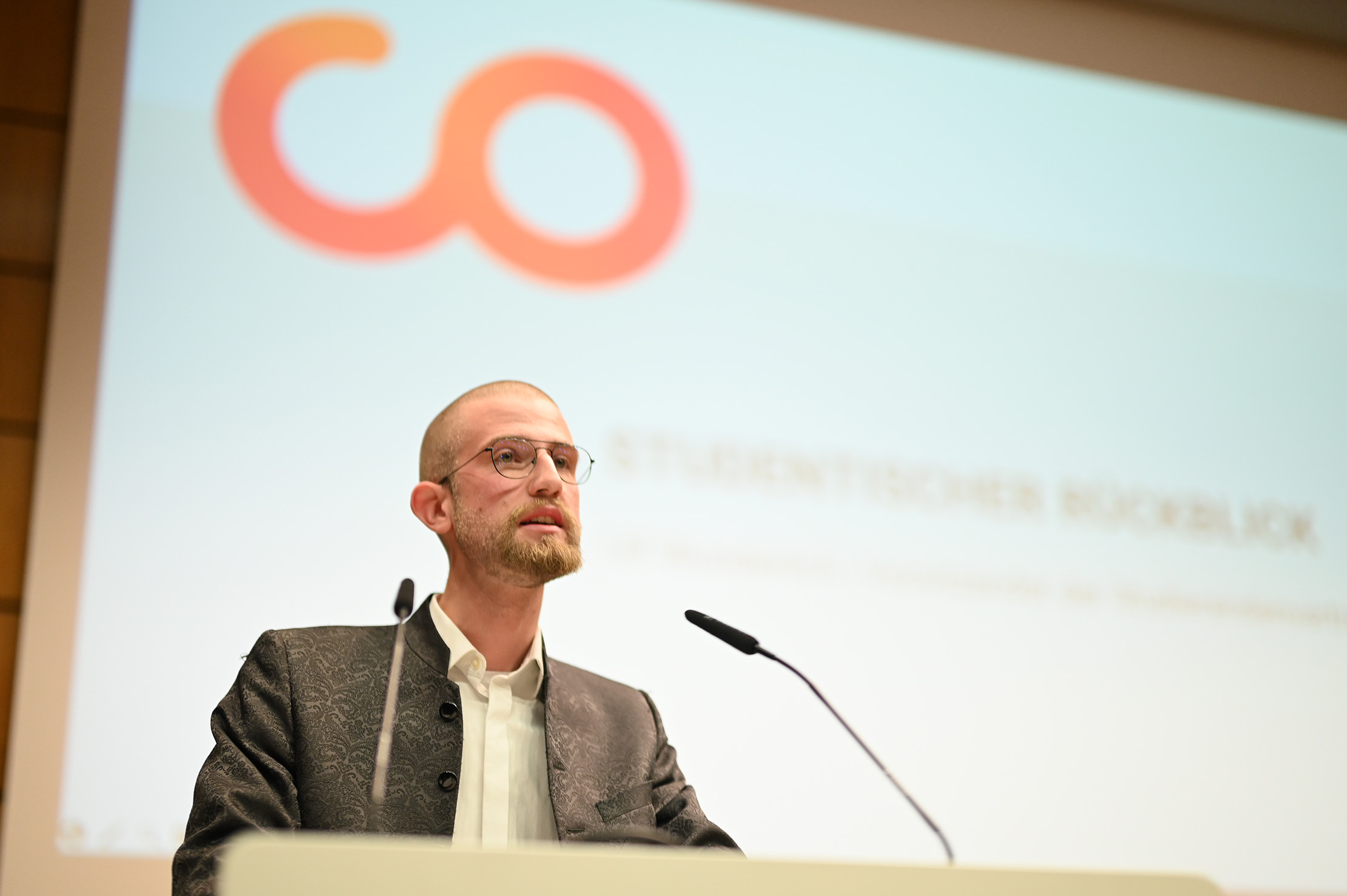 Eine Person mit Brille und Bart spricht an einem Podium vor einem verschwommenen Präsentationsbildschirm. Auf dem Bildschirm sind ein orange-rotes Logo zu sehen, das den Buchstaben „co“ ähnelt, und darunter ein Teiltext.