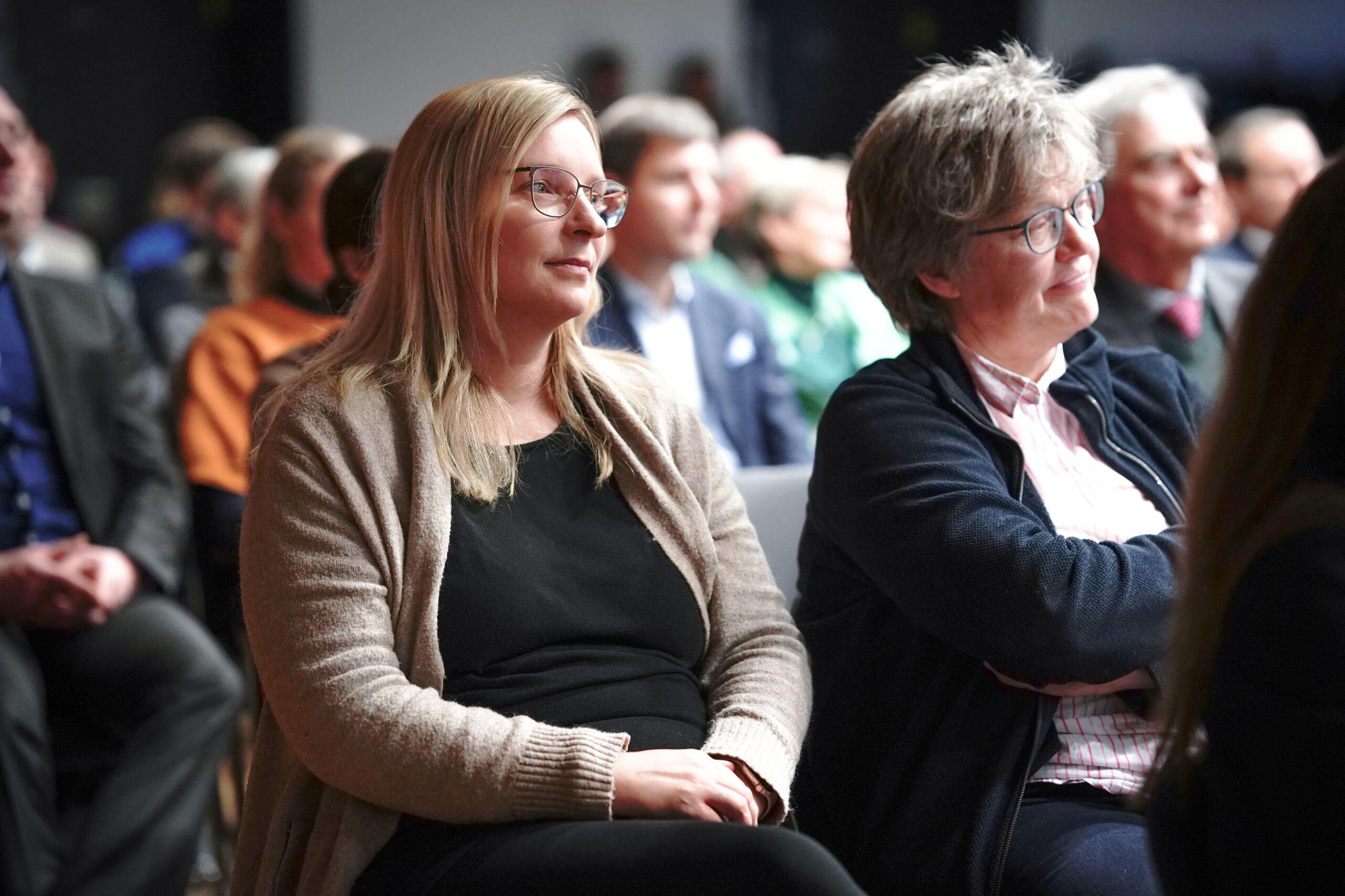 Eine Gruppe von Menschen sitzt in einer Konferenzatmosphäre und hört aufmerksam zu. Zwei Frauen im Vordergrund, eine mit Brille und beiger Strickjacke, die andere mit kurzen Haaren und Brille, fokussieren auf den Vortragenden.