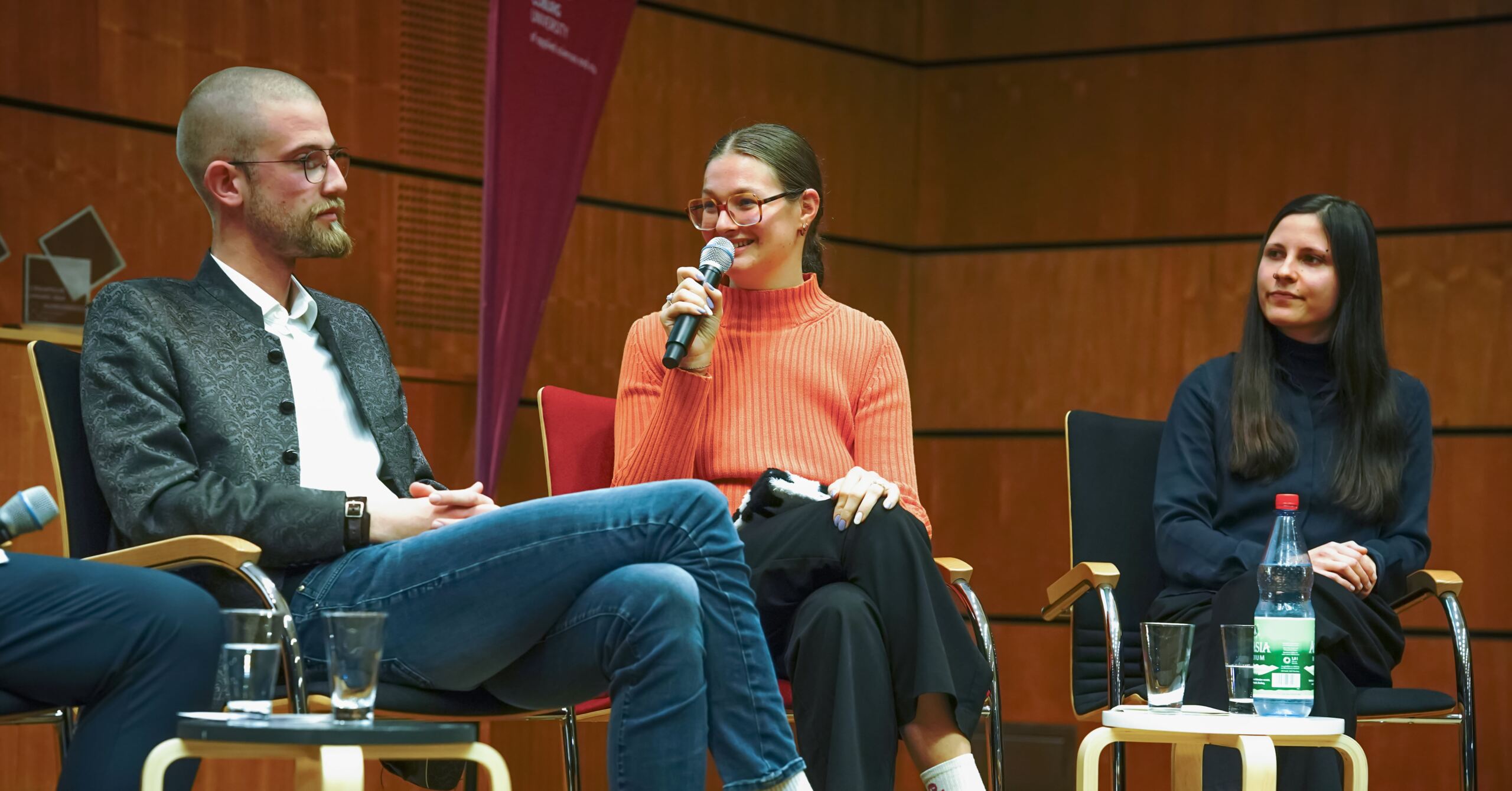 Drei Personen sitzen in einem Panel. Die Person in der Mitte trägt eine Brille und einen orangefarbenen Pullover und hält ein Mikrofon. Die anderen beiden hören aufmerksam zu. Sie sitzen in einem modernen Raum mit holzgetäfelten Wänden.