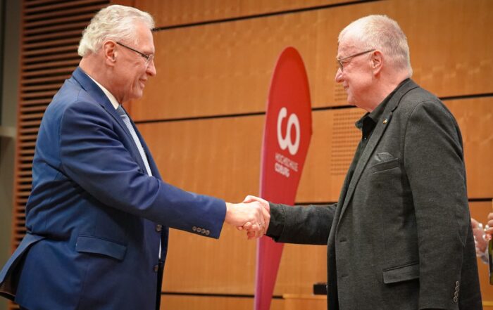 Zwei Männer geben sich in einem formellen Rahmen die Hand. Der Mann links trägt einen blauen Anzug und der Mann rechts einen dunklen Blazer. Sie stehen vor einem rot-weißen Banner mit dem Wort „GO“ im Vordergrund. Der Hintergrund ist mit Holzvertäfelung versehen.