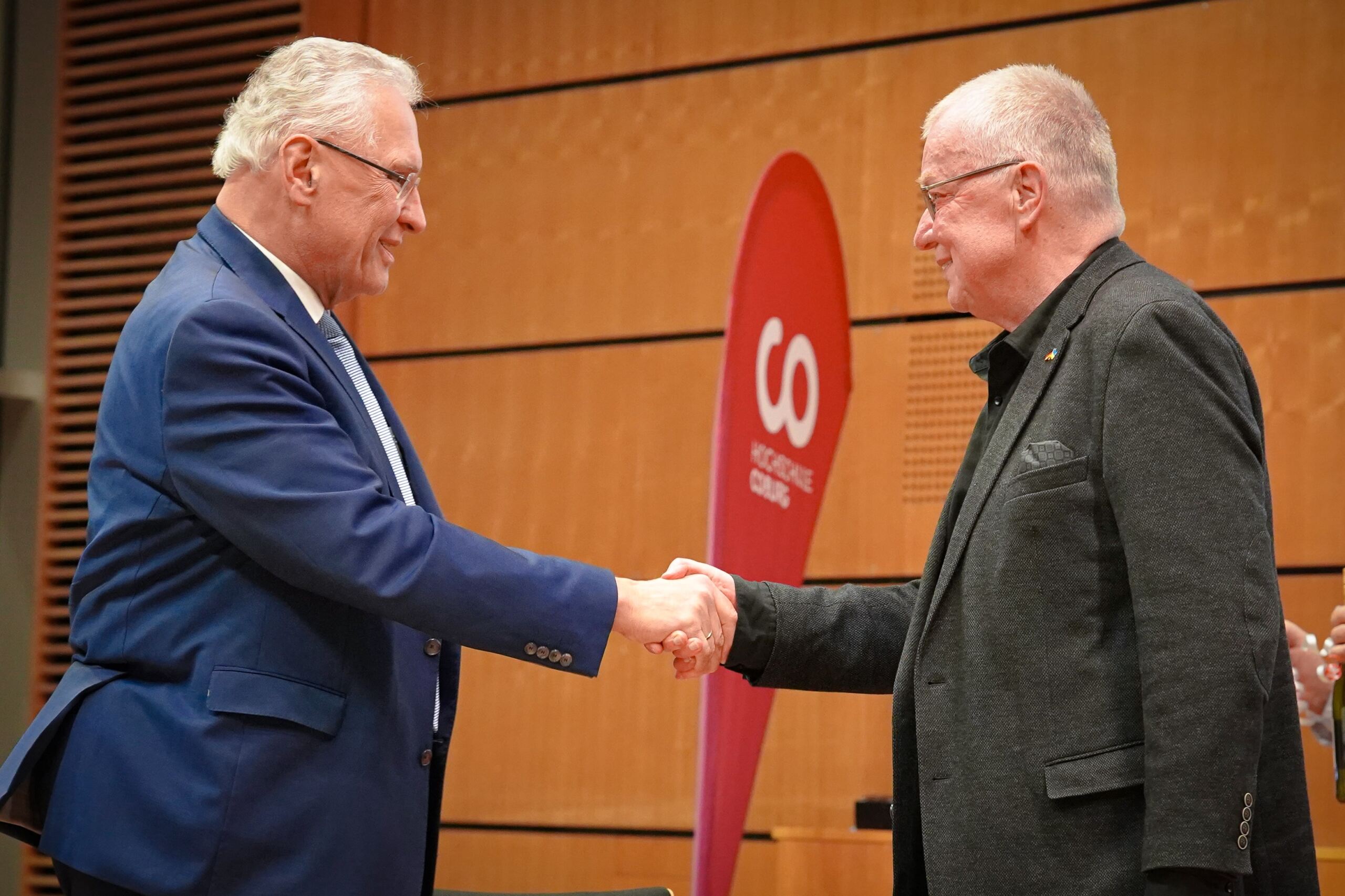 Zwei Männer geben sich in einem formellen Rahmen die Hand. Der Mann links trägt einen blauen Anzug und der Mann rechts einen dunklen Blazer. Sie stehen vor einem rot-weißen Banner mit dem Wort „GO“ im Vordergrund. Der Hintergrund ist mit Holzvertäfelung versehen.