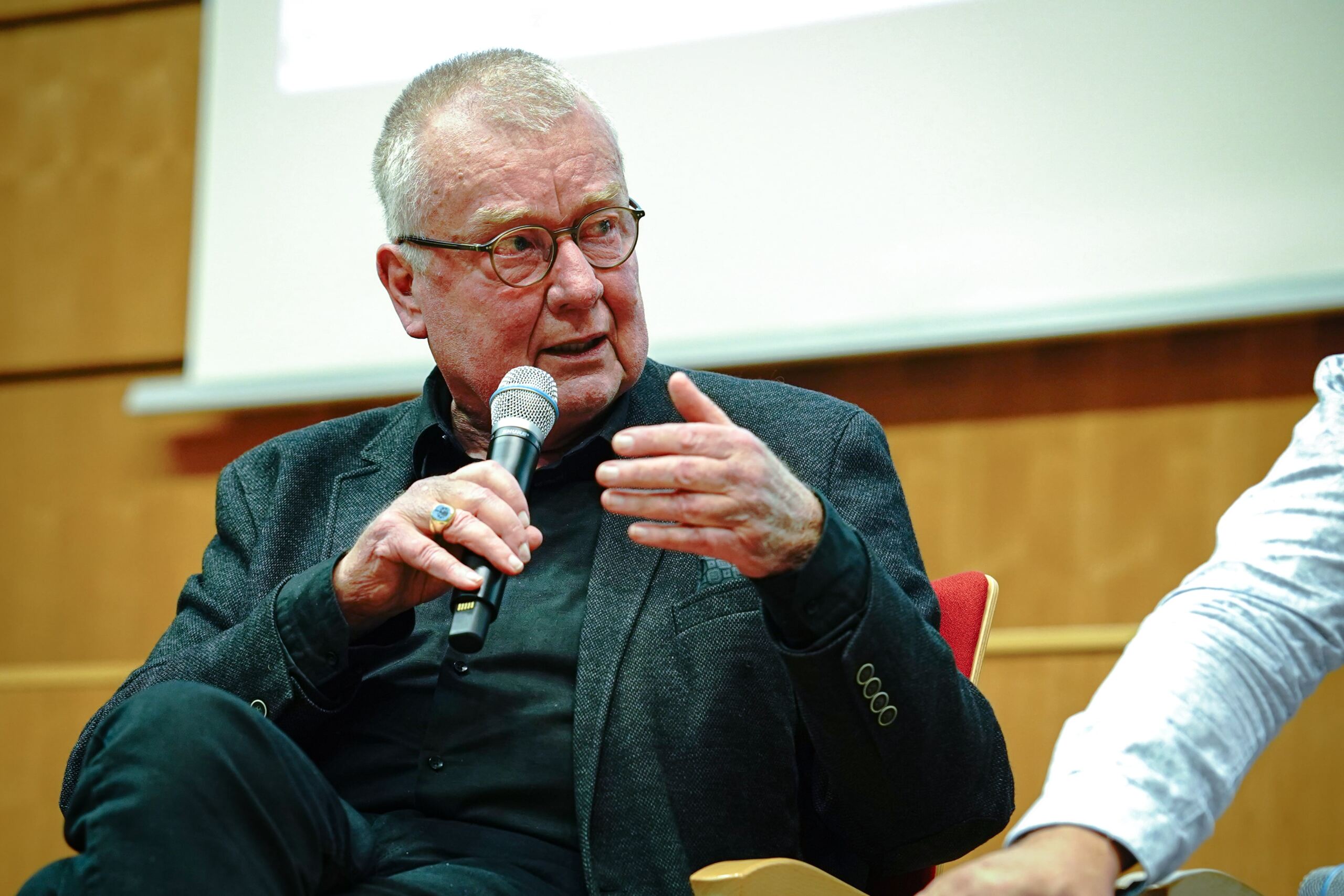 Ein älterer Mann mit Brille spricht im Sitzen in ein Mikrofon. Er trägt ein schwarzes Hemd und einen dunklen Blazer und gestikuliert mit der linken Hand. Der Hintergrund ist eine Holzplatte mit einem weißen Abschnitt am oberen Rand.