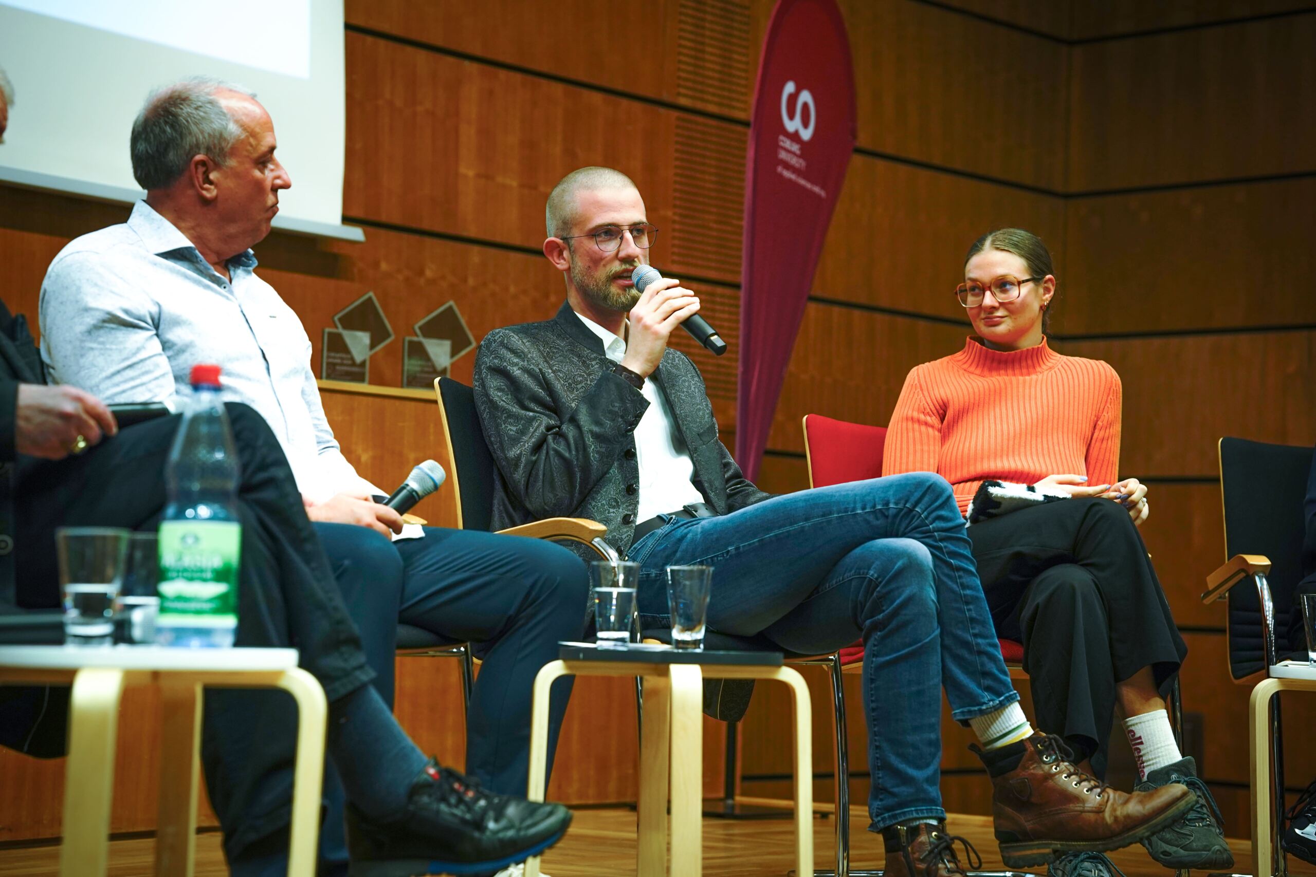 Drei Personen sitzen in einem Konferenzraum an einem Panel. Der Mann in der Mitte hält ein Mikrofon und spricht. Rechts von ihm sitzt eine Frau in einem orangefarbenen Pullover. Auf dem Tisch vor ihnen stehen Mikrofone und Wasserflaschen.