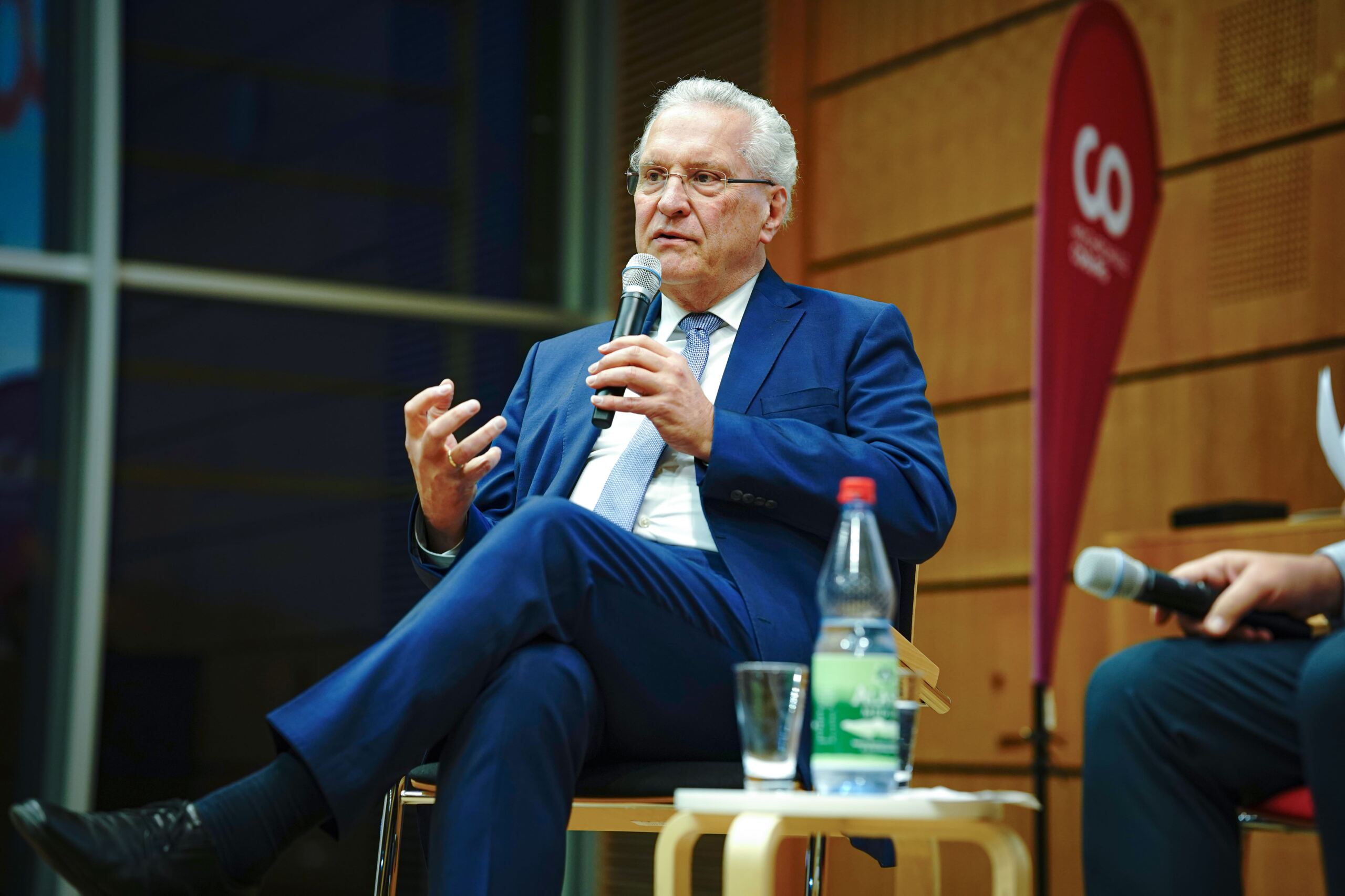 Ein älterer Mann im blauen Anzug sitzt auf der Bühne, hält ein Mikrofon und gestikuliert mit der Hand. Im Hintergrund ist ein rot-weißes Banner zu sehen. Vor ihm steht ein kleiner Tisch mit einer Flasche Wasser und einem Glas.