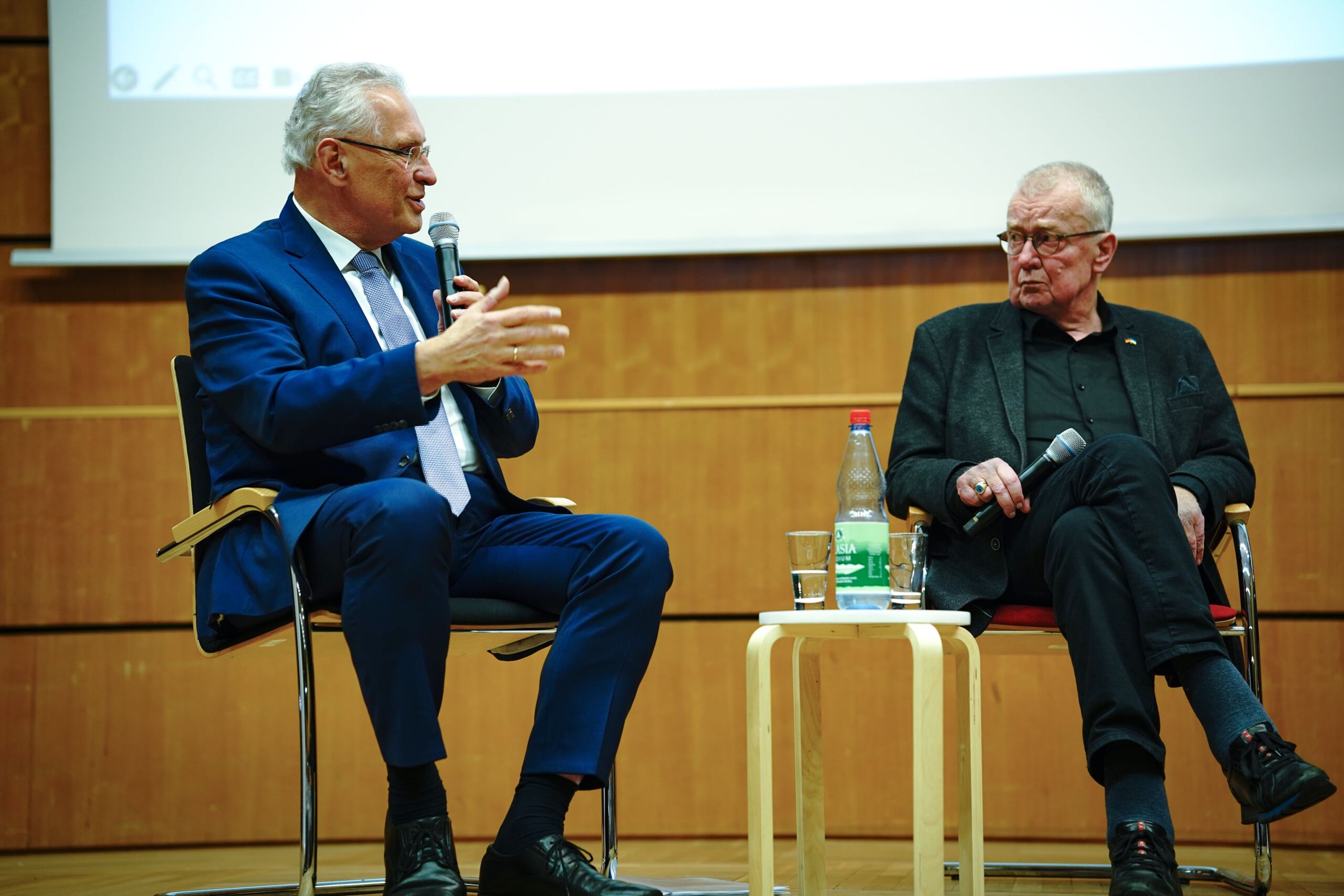 Auf der Bühne sitzen zwei Männer und diskutieren. Einer trägt einen blauen Anzug und gestikuliert mit der Hand, während er in ein Mikrofon spricht. Der andere, in einer schwarzen Jacke, hält ein Mikrofon und hört aufmerksam zu. Zwischen ihnen steht ein kleiner Tisch mit einer Wasserflasche.