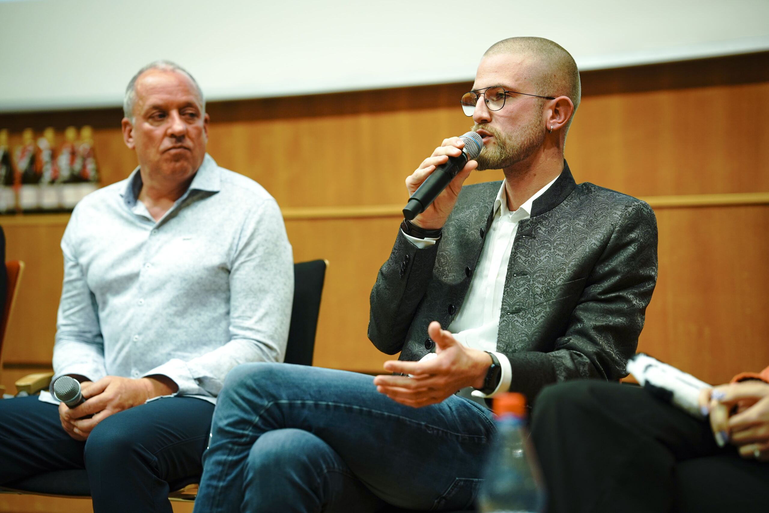 Zwei Männer sitzen bei einer Podiumsdiskussion. Der rechte Mann, mit Brille und gemustertem Blazer, spricht in ein Mikrofon und gestikuliert dabei mit der Hand. Der linke Mann, im hellen Hemd, hört aufmerksam zu. Beide stehen auf einer Bühne mit braunem Hintergrund.