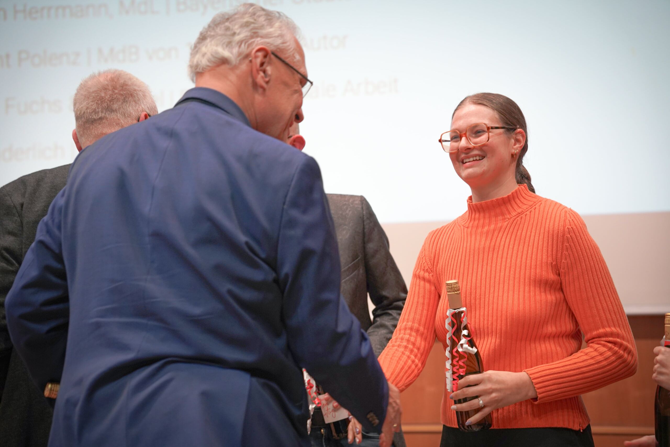 Eine Frau in einem leuchtend orangefarbenen Pullover lächelt und schüttelt einem älteren Mann in einem blauen Blazer die Hand. Sie hält eine eingewickelte Flasche in der Hand. Sie befinden sich in einem Raum mit einer Projektionsleinwand im Hintergrund. Mehrere andere Personen sind in der Nähe.