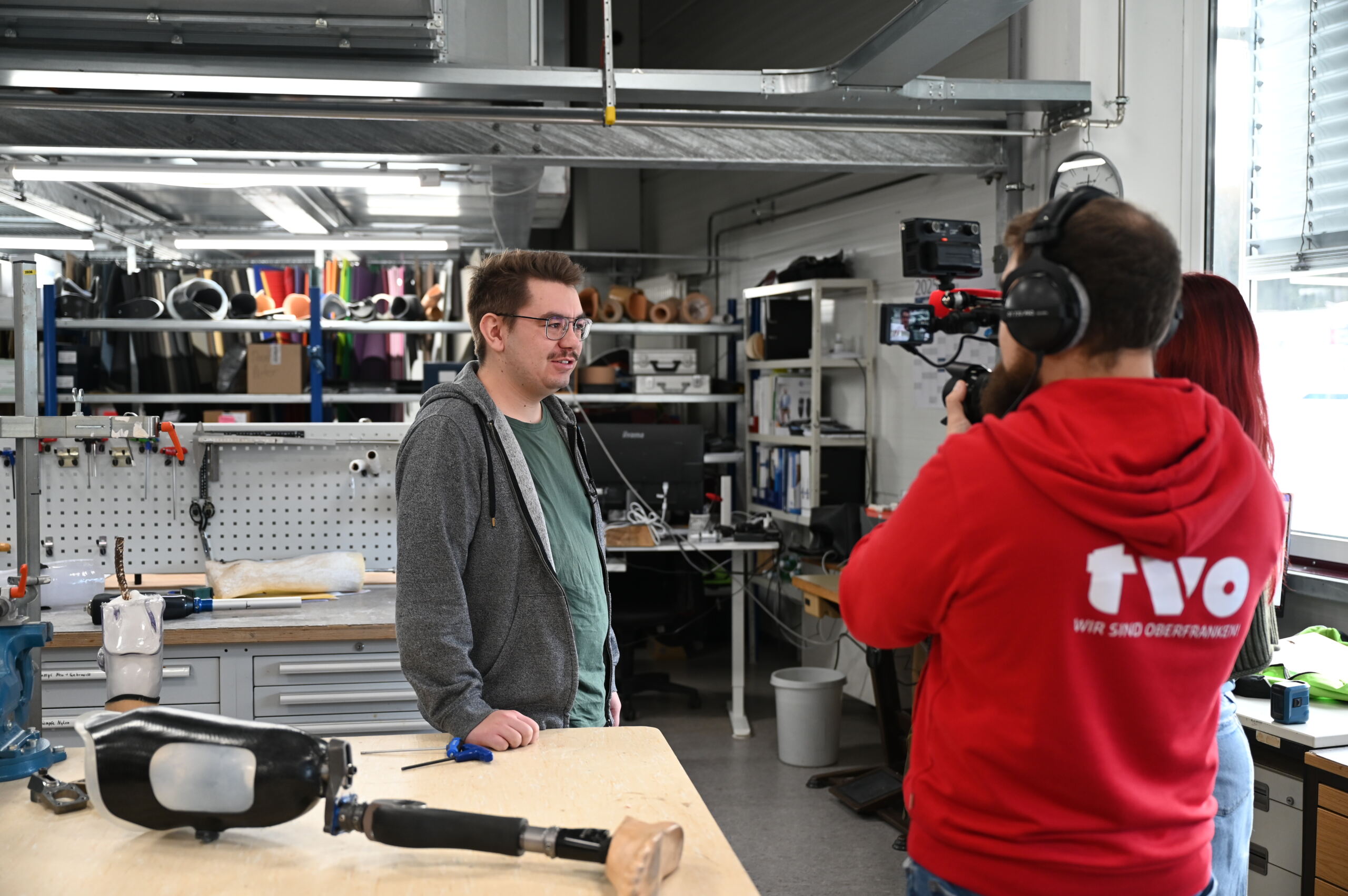 Eine Person in Freizeitkleidung wird in einer Werkstatt von einem zweiköpfigen Kamerateam gefilmt. Im Vordergrund liegt eine Beinprothese auf dem Tisch. Die Werkstatt ist voll mit Werkzeugen, Materialien und Geräten.