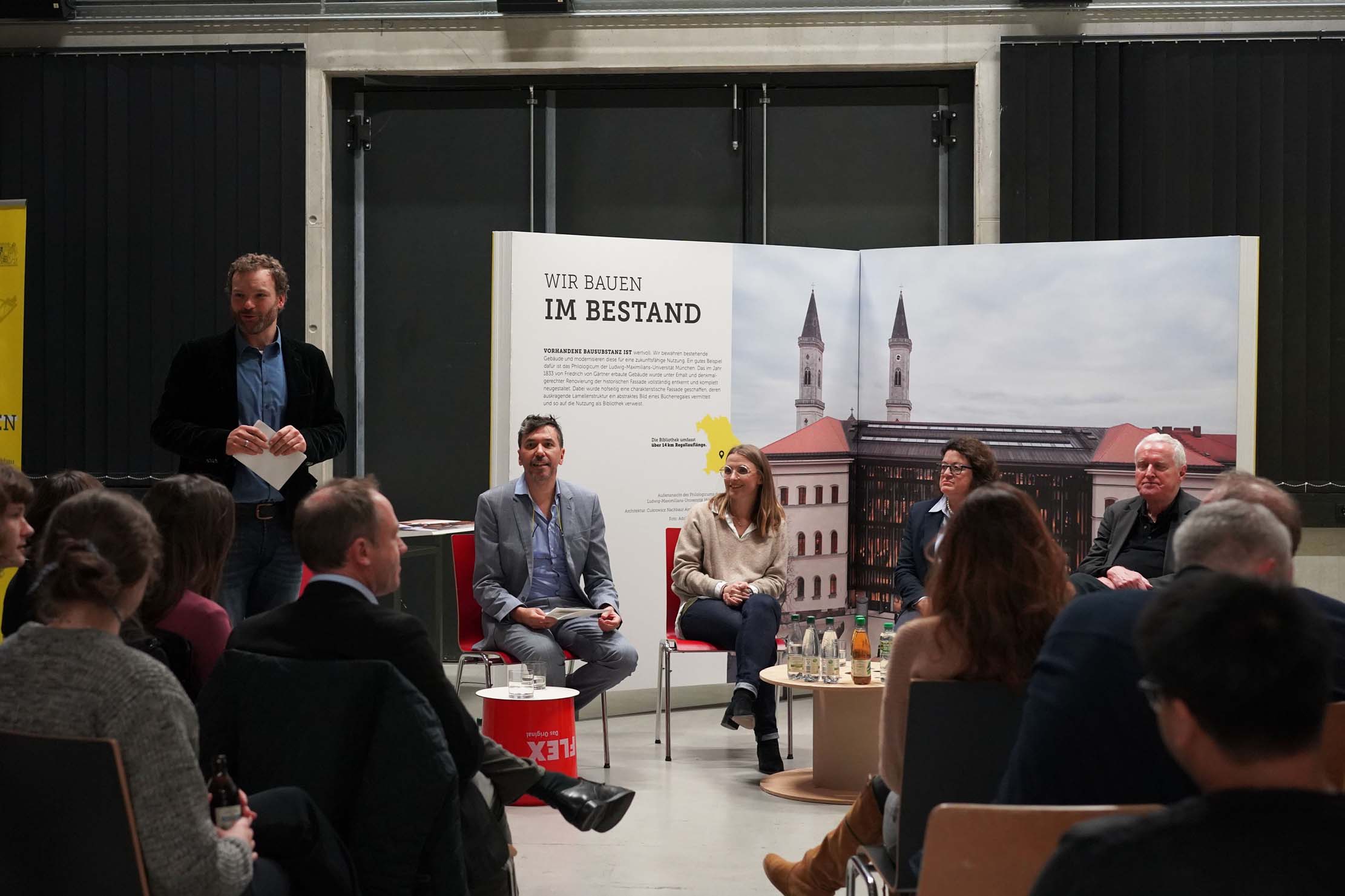 Podiumsdiskussion mit fünf Personen auf der Bühne. Eine Person steht auf und wendet sich an das Publikum. Dahinter hängt ein Plakat mit dem Text „Wir bauen im Bestand“ und einem Bild eines Gebäudes. Das Publikum hört aufmerksam zu.