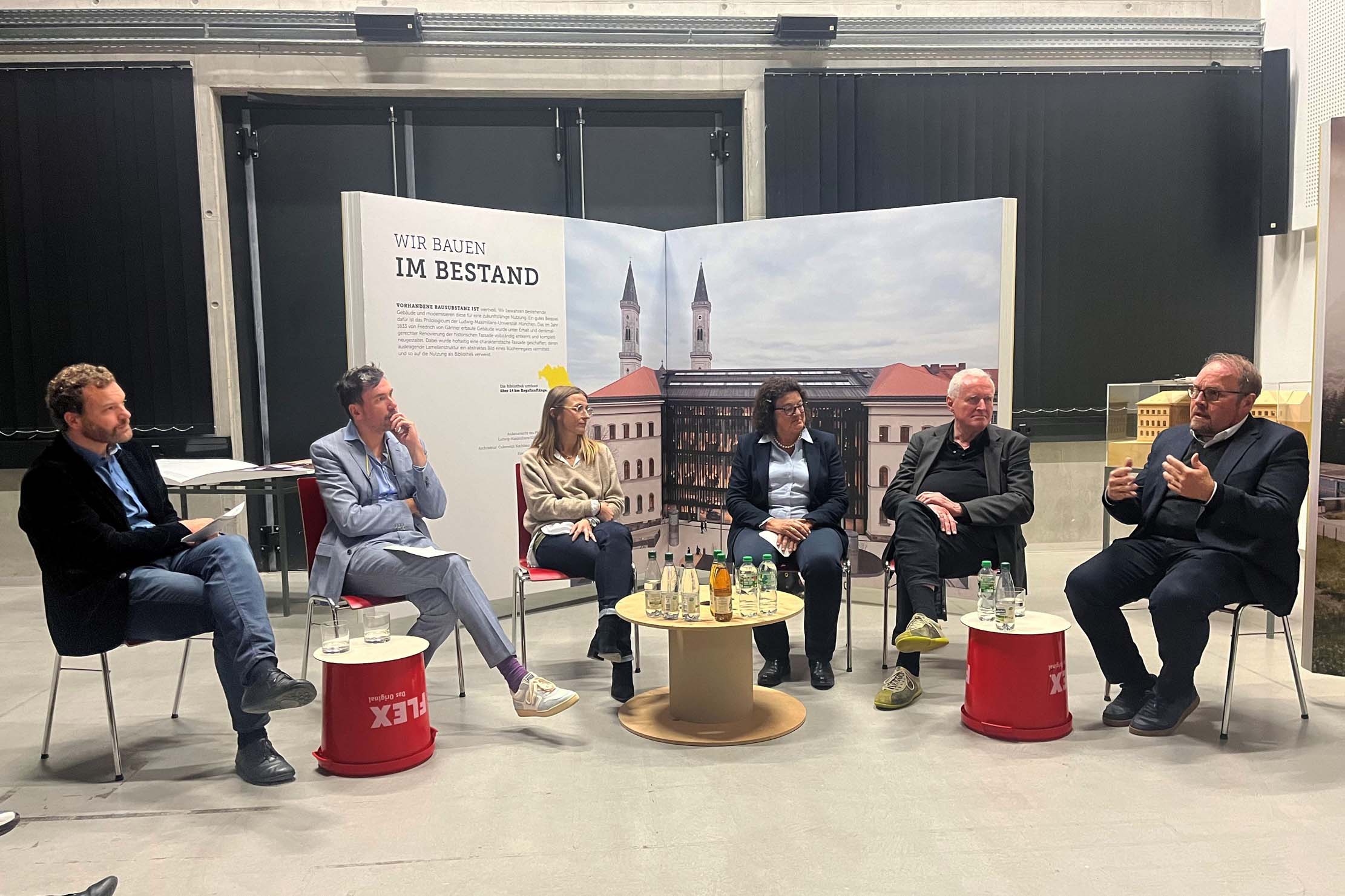 Ein Panel aus sechs Personen sitzt im Halbkreis auf Stühlen und diskutiert. Dahinter ist eine Rückwand mit einem Architekturbild und einem Text in deutscher Sprache zu sehen. Vor ihnen stehen auf kleinen Tischen verschiedene Getränke.