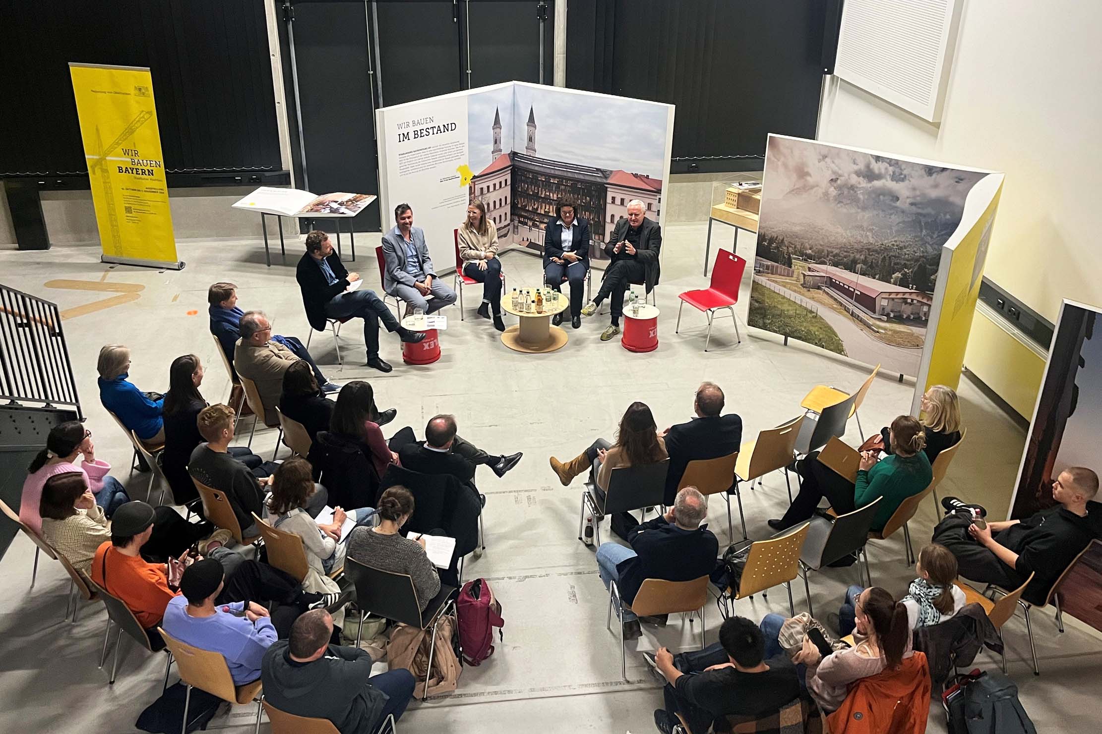 Eine Gruppe von Menschen sitzt während einer Podiumsdiskussion in einem großen Raum im Kreis. Das Podium besteht aus vier Personen auf der Bühne, während das Publikum aufmerksam zuhört. Hinter dem Podium hängen große Poster mit Gebäudeabbildungen.