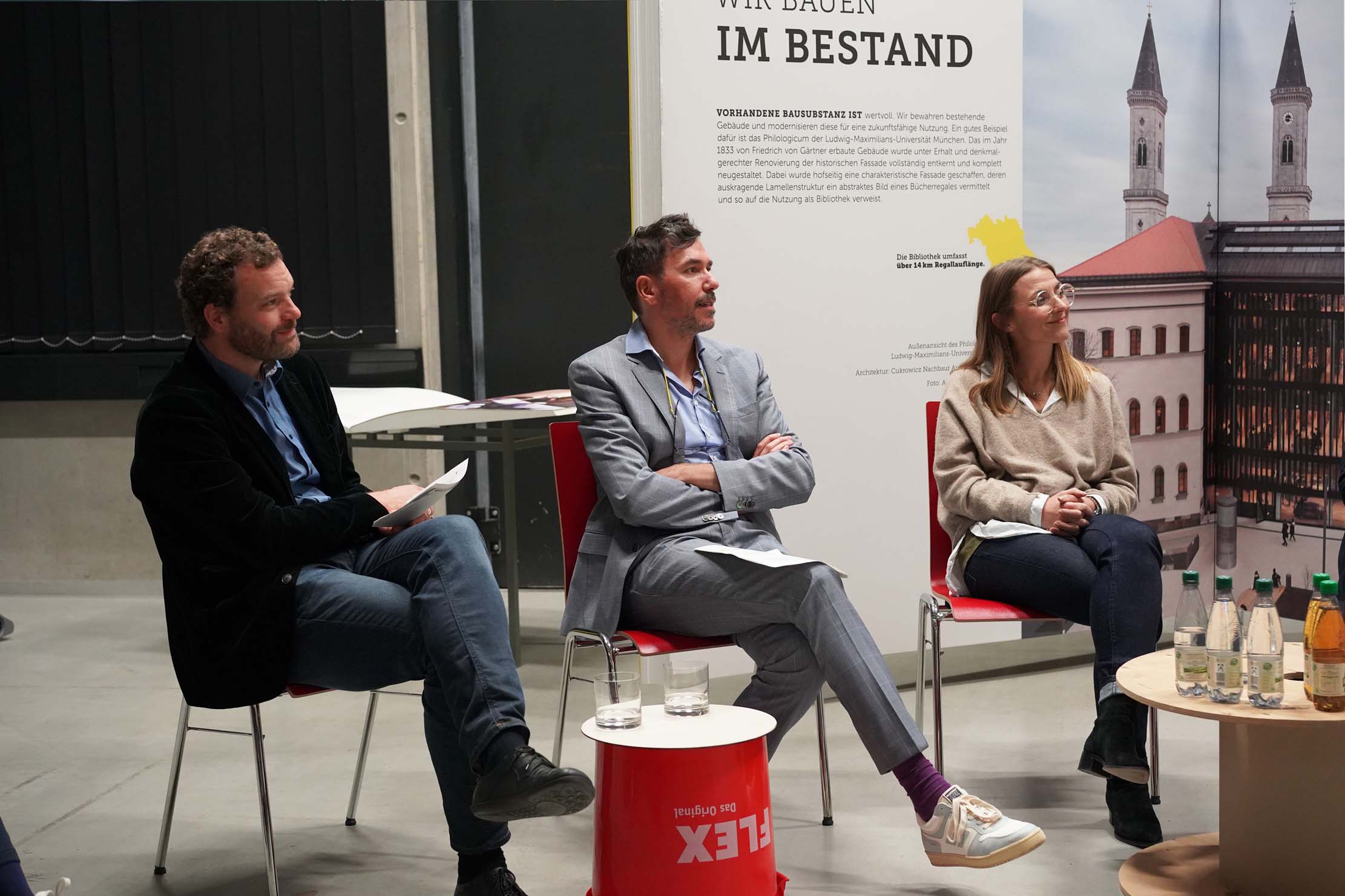 Drei Personen sitzen während einer Podiumsdiskussion auf Stühlen. Sie befinden sich in einem modernen Raum mit einem Poster und Architekturbildern im Hintergrund. Flaschen und Papiere liegen auf einem kleinen runden Tisch in der Nähe.