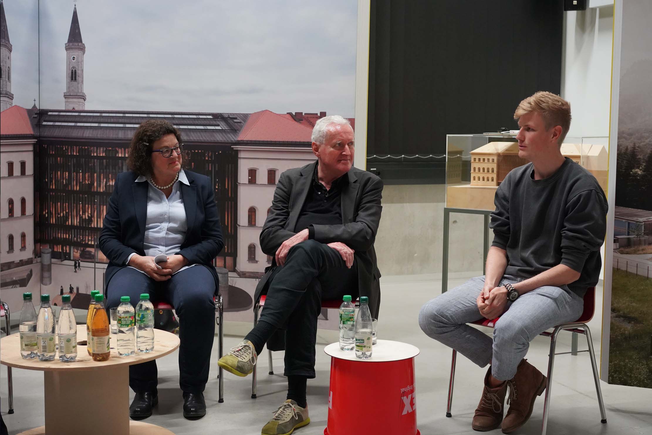 Drei Personen sitzen in einer Podiumsdiskussionsanordnung. Sie befinden sich in einem modernen Raum mit einem großen Gebäude im Hintergrund. Auf einem Tisch davor stehen Getränkeflaschen. Die Personen unterhalten sich, was auf eine spannende Diskussion schließen lässt.