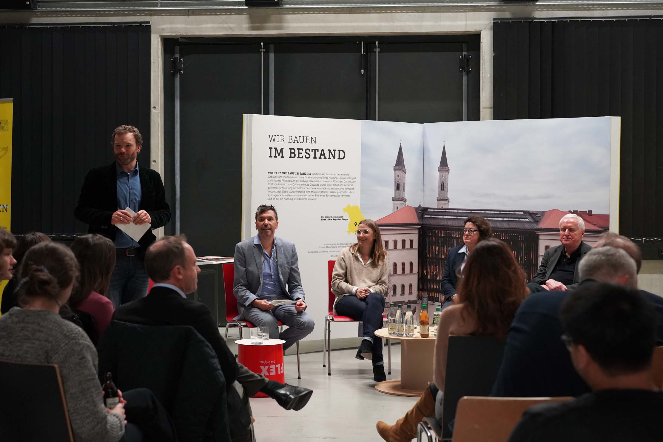 Eine Gruppe von fünf Personen, drei sitzend und zwei stehend, diskutieren in einem Raum mit Publikum. Hinter ihnen ist ein großes Plakat mit einer Stadtansicht und dem deutschen Text „Wir bauen im Bestand“ zu sehen. Flaschengetränke stehen auf einem Tisch vor den Diskussionsteilnehmern.