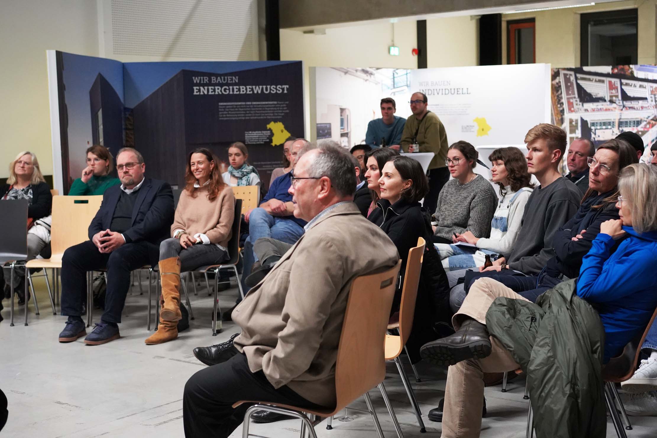 Eine Gruppe von Menschen sitzt im Halbkreis und hört einem Vortragenden in einem modernen Innenraum aufmerksam zu. Im Hintergrund sind Plakate und Informationstafeln zu sehen.