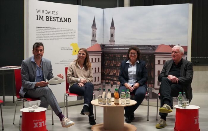 Vier Personen sitzen bei einer Podiumsdiskussion. Hinter ihnen ist ein Display mit Architekturbildern und Text zu sehen. Auf dem Tisch vor ihnen stehen Wasserflaschen und Getränke. Sie sitzen auf roten Stühlen vor schwarzem Hintergrund.