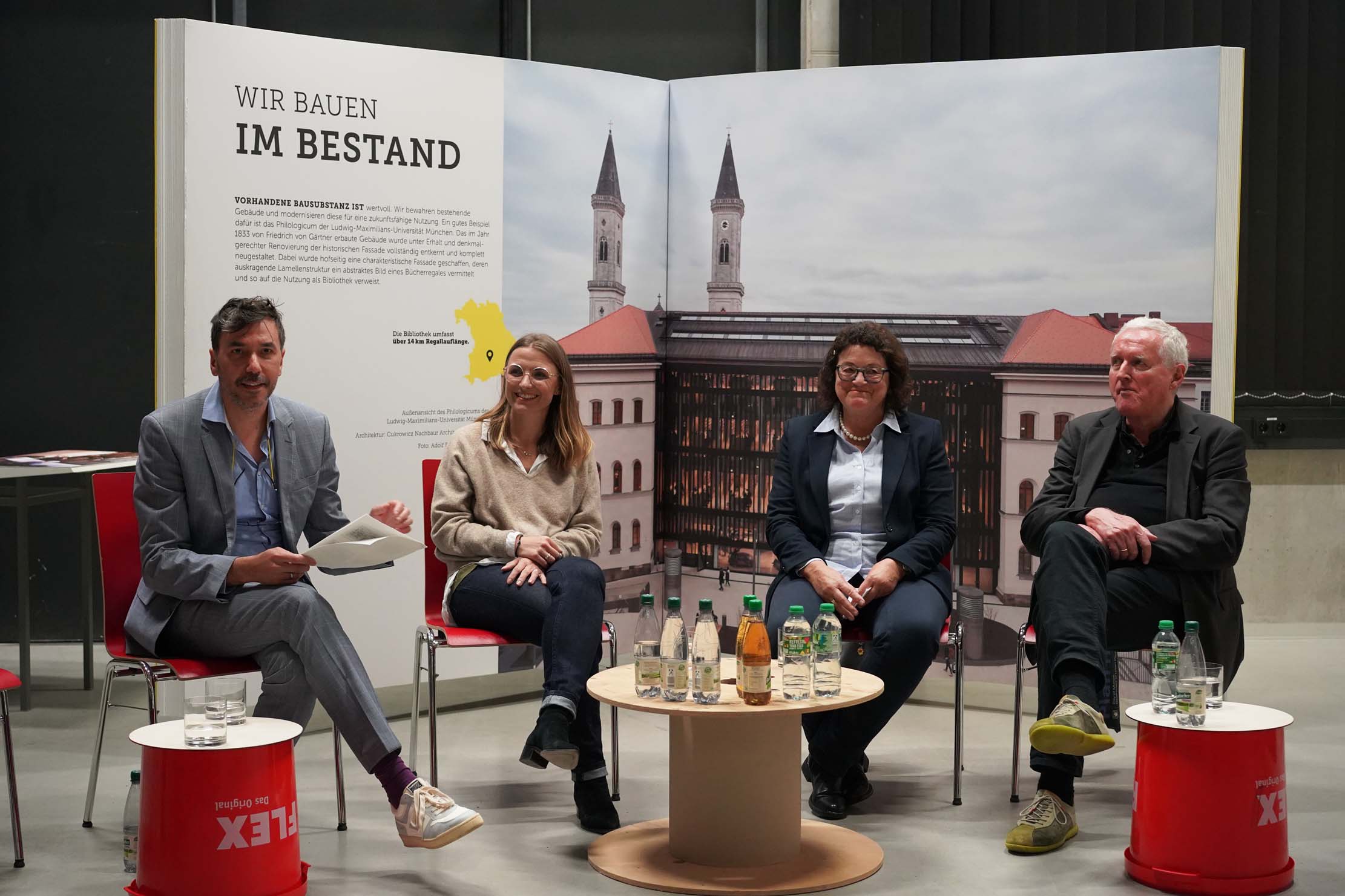 Vier Personen sitzen bei einer Podiumsdiskussion. Hinter ihnen ist ein Display mit Architekturbildern und Text zu sehen. Auf dem Tisch vor ihnen stehen Wasserflaschen und Getränke. Sie sitzen auf roten Stühlen vor schwarzem Hintergrund.