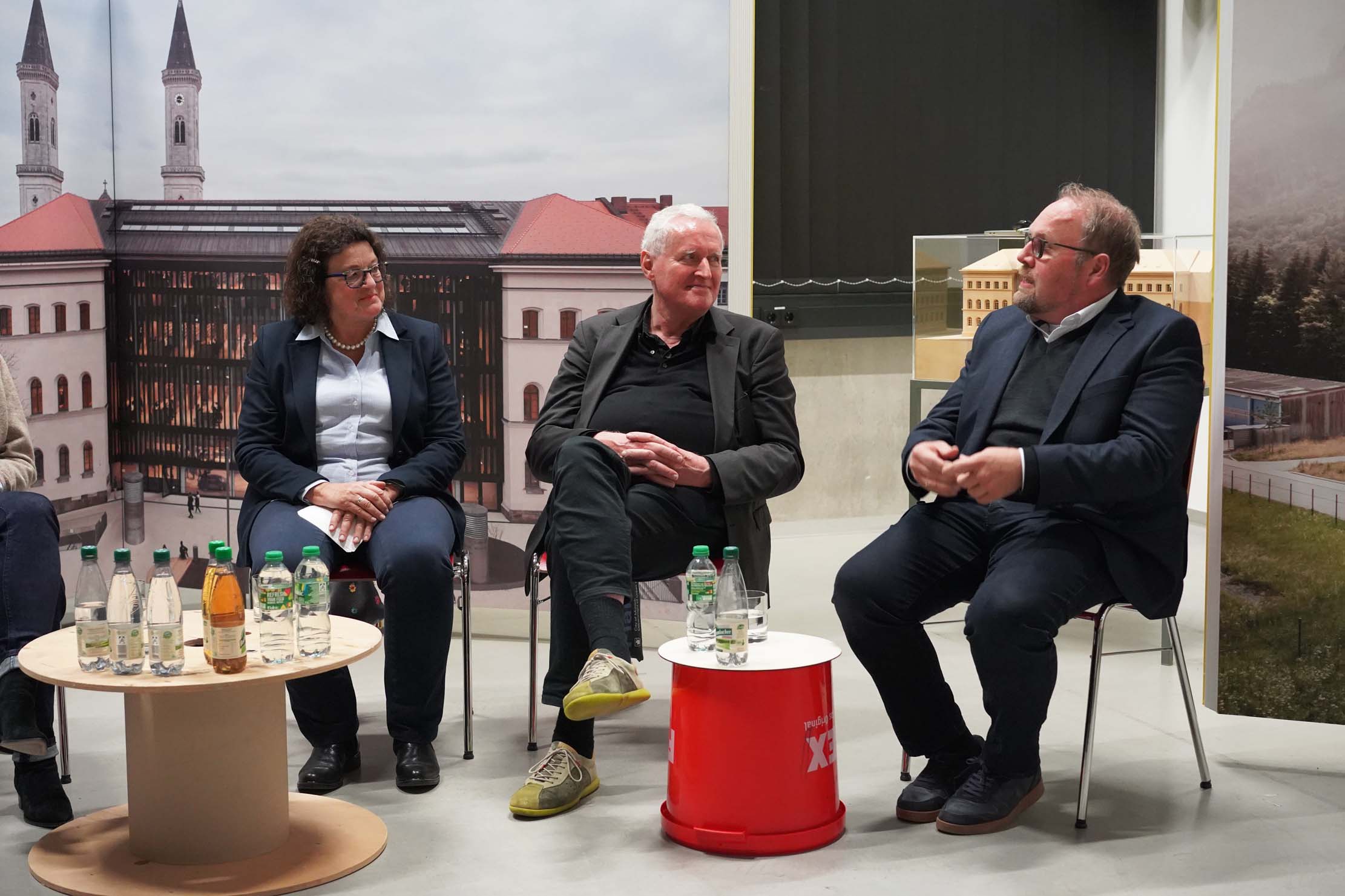 Drei Personen sitzen bei einer Podiumsdiskussion. Zwei Männer und eine Frau unterhalten sich vor der Kulisse eines Gebäudes und von Türmen. Flaschen mit Wasser und Tee stehen auf einem kleinen Tisch. Die Atmosphäre wirkt professionell und leger.